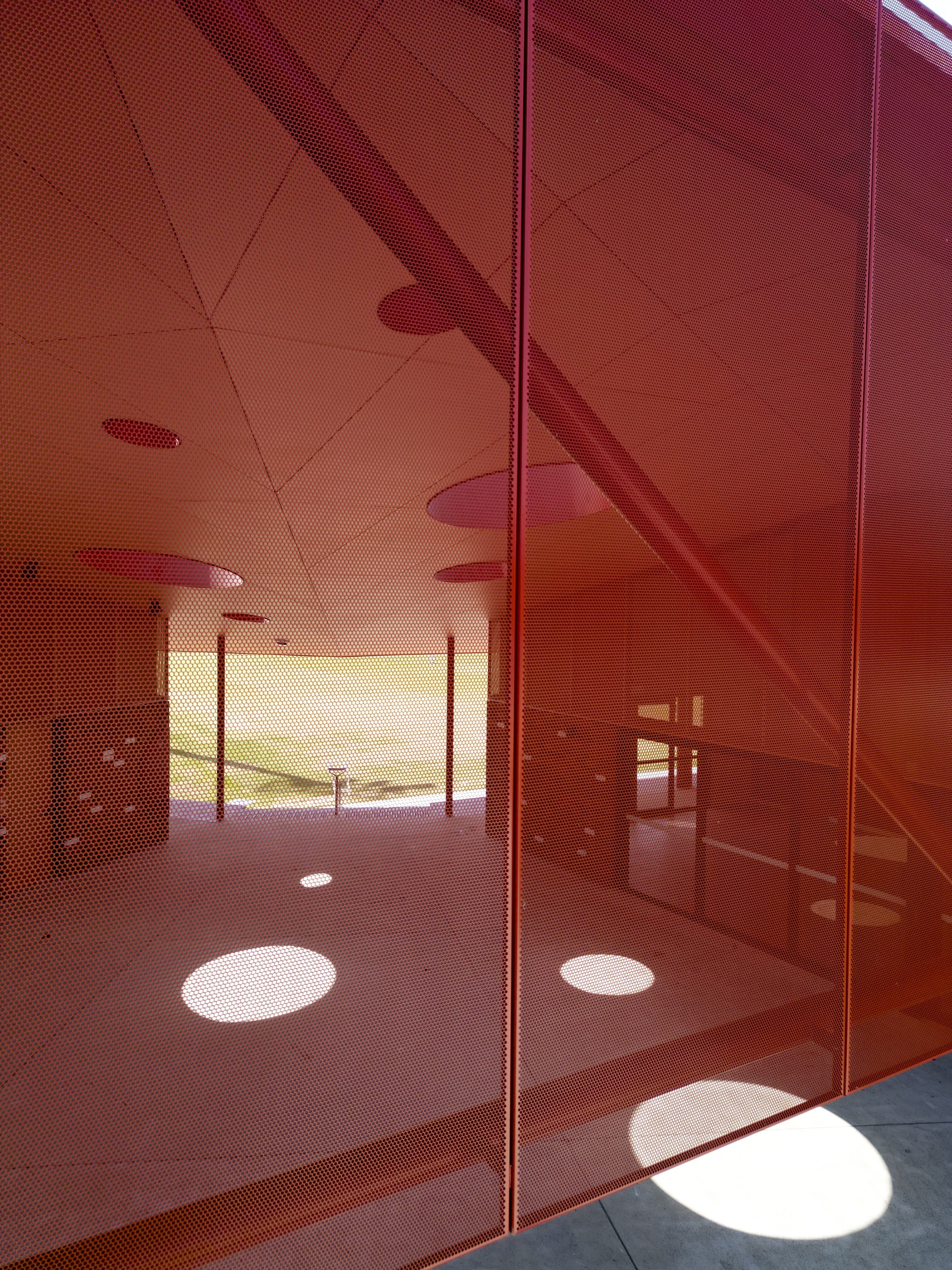 Reflections of circular ceiling apertures and columns on red perforated metal panels in afternoon sunlight