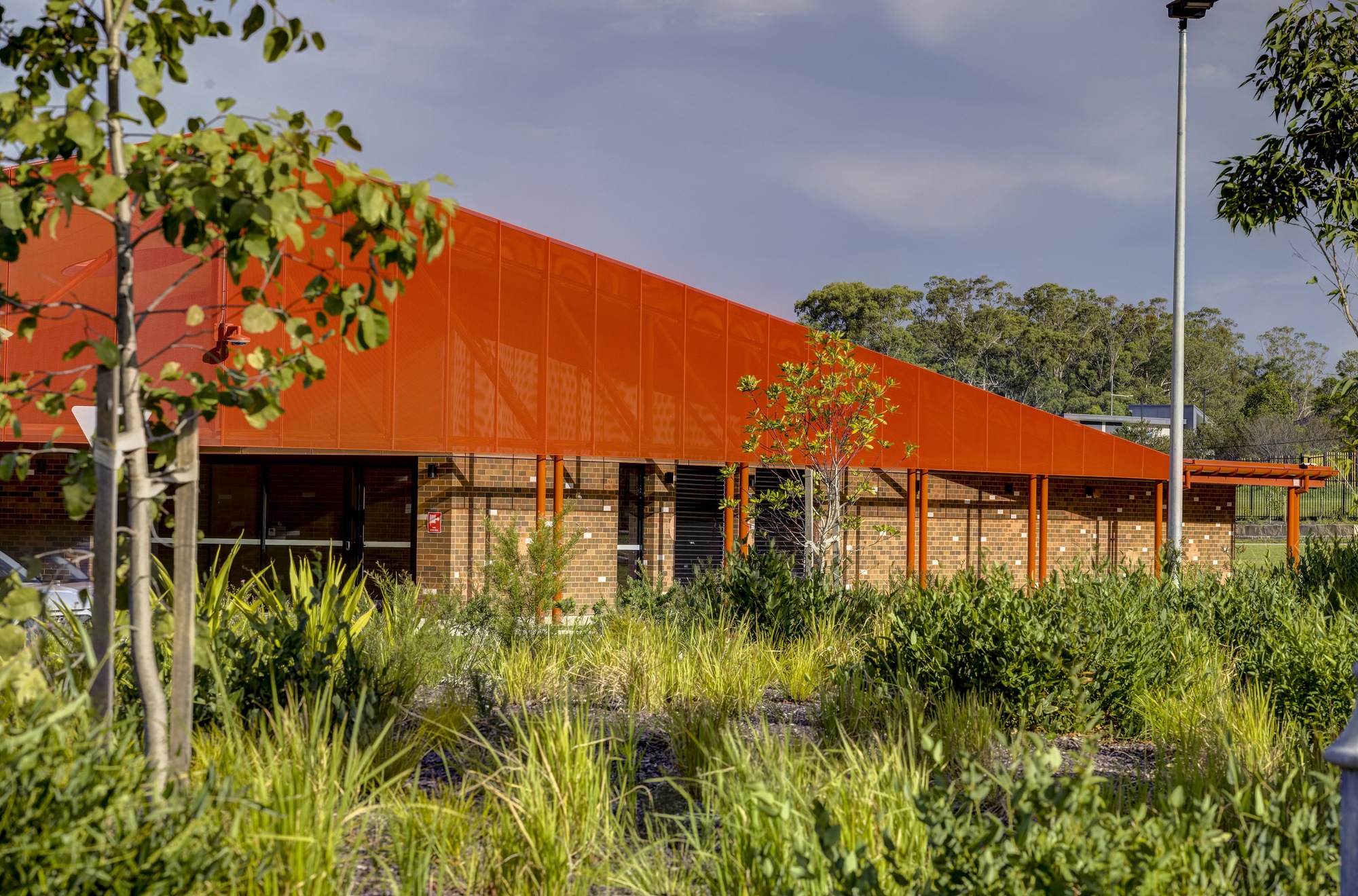 Long sloped orange metal canopy over brick walls viewed across native grasses and young trees