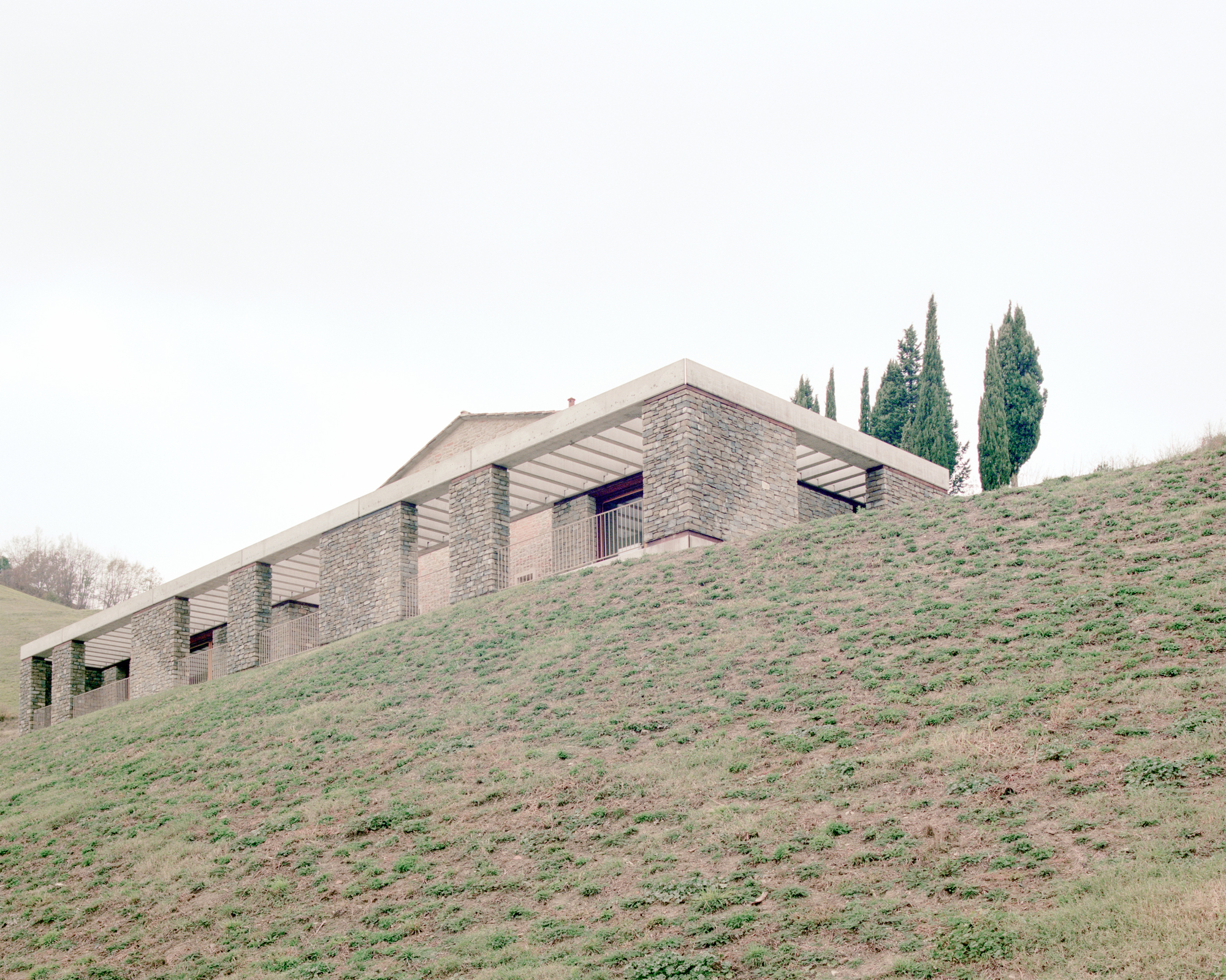 Stone-clad residence emerging from a grassy hillside with cypress trees under an overcast sky