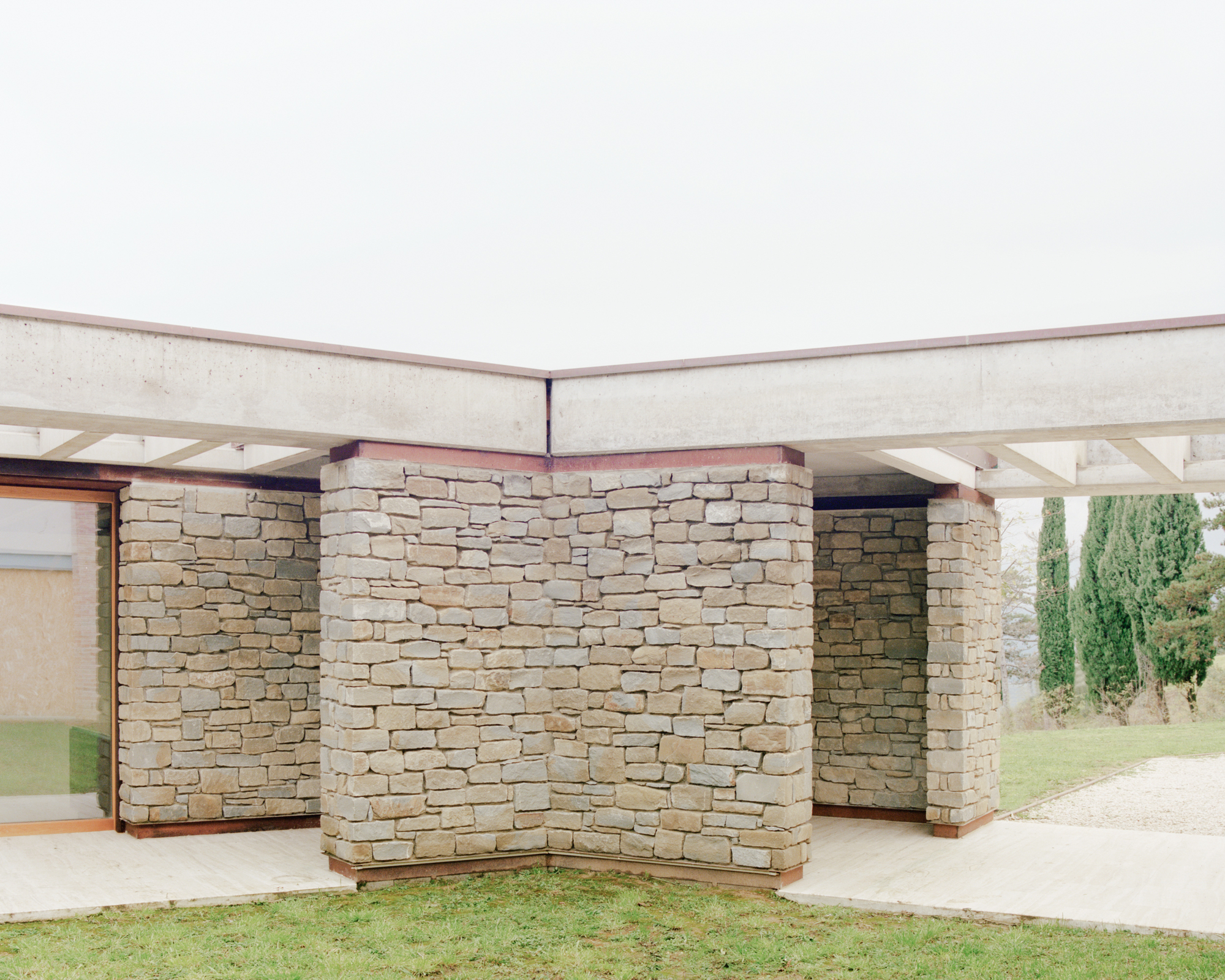 Layered stone walls beneath a flat concrete overhang with a lawn and cypress trees in the background