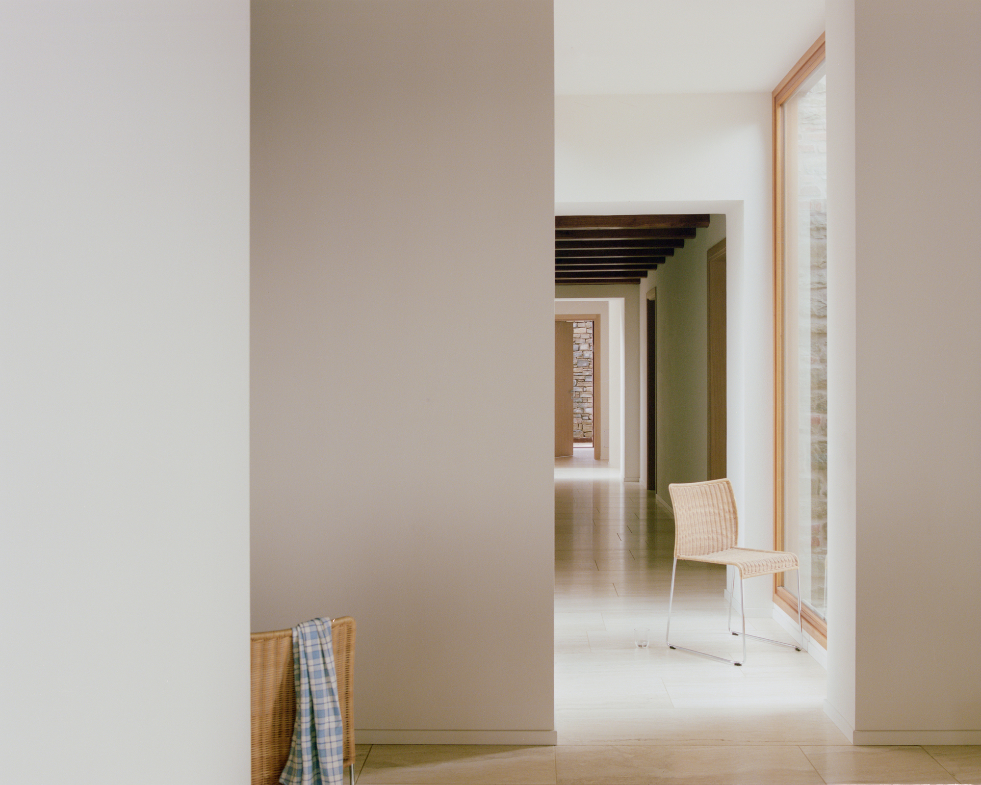 Corridor view through white walls and doorways revealing stone column and wicker chair in daylight