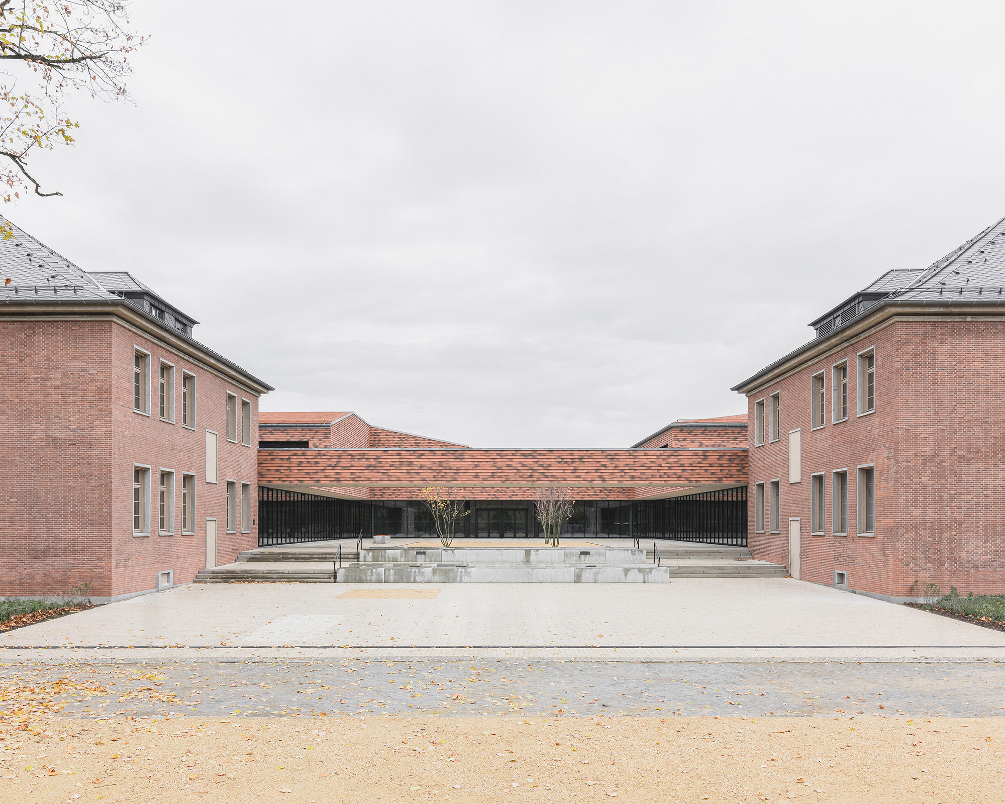 Symmetrical courtyard framed by pink brick volumes with a curved roof linking two wings under an overcast sky