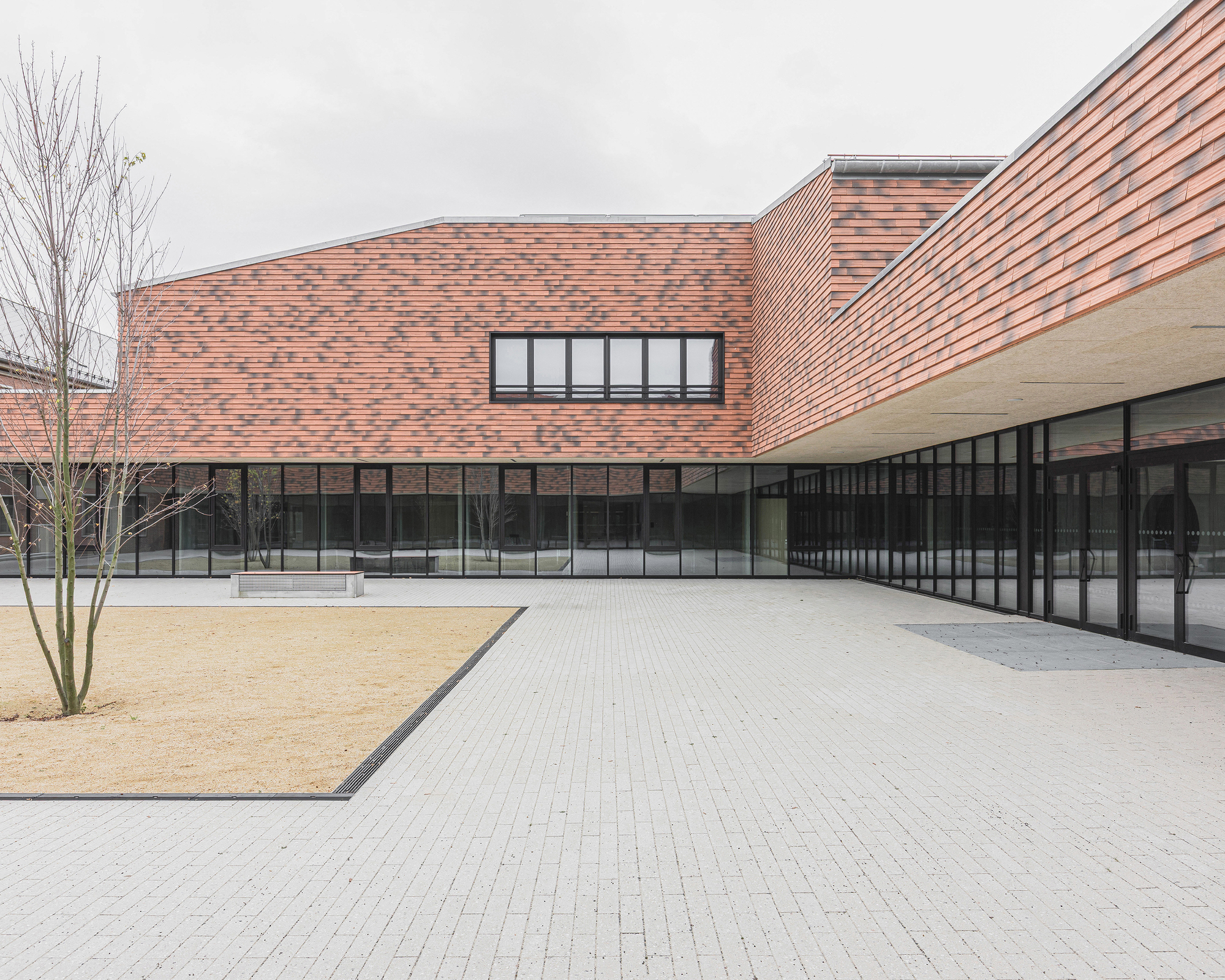 Paved courtyard with gravel bed and bare trees beneath pink brick facades and black-framed glazing