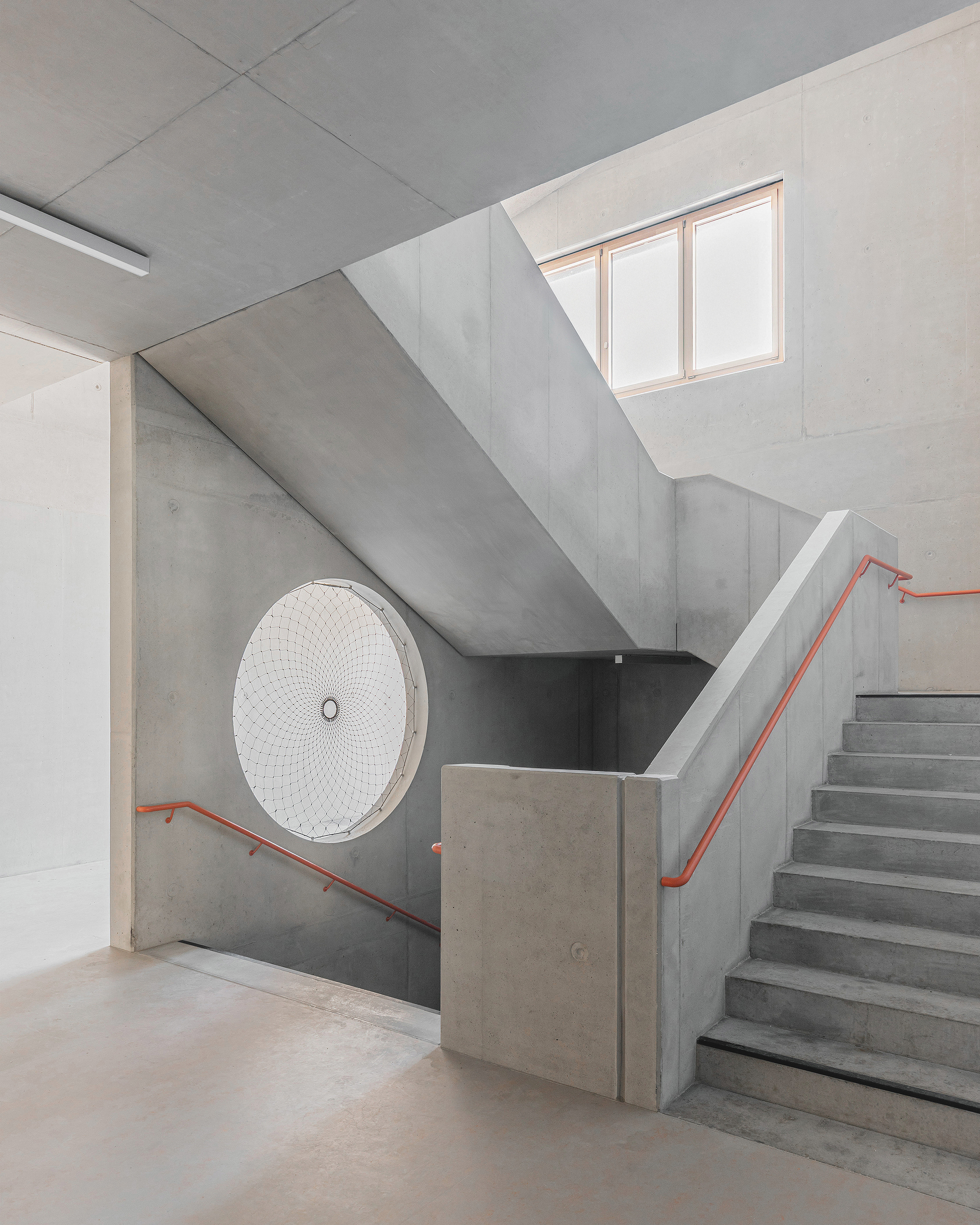Concrete stairwell with orange handrails ascending toward a skylight above a circular decorative grille