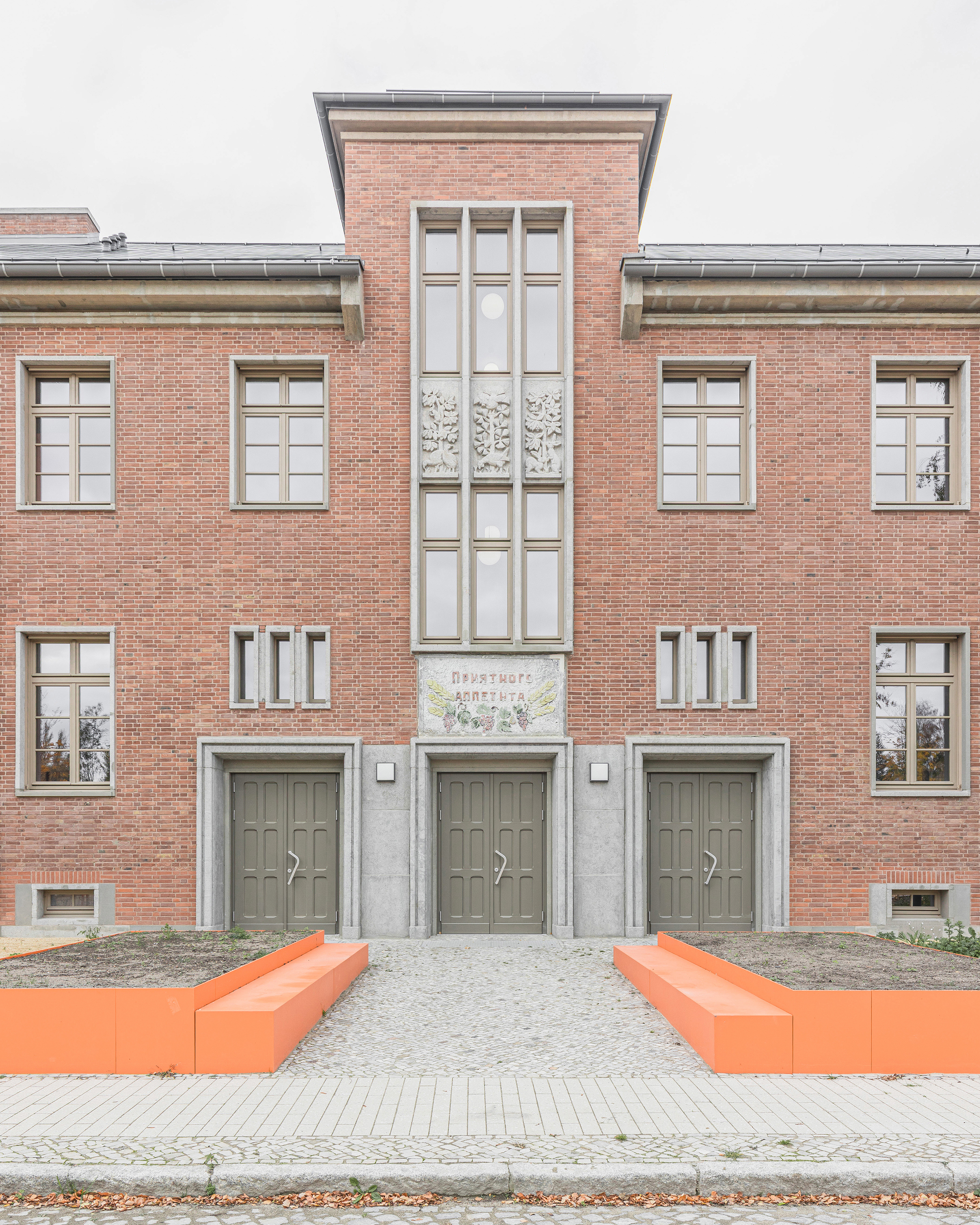 Red brick facade with tall glazed bay and orange benches flanking the entrance under overcast sky
