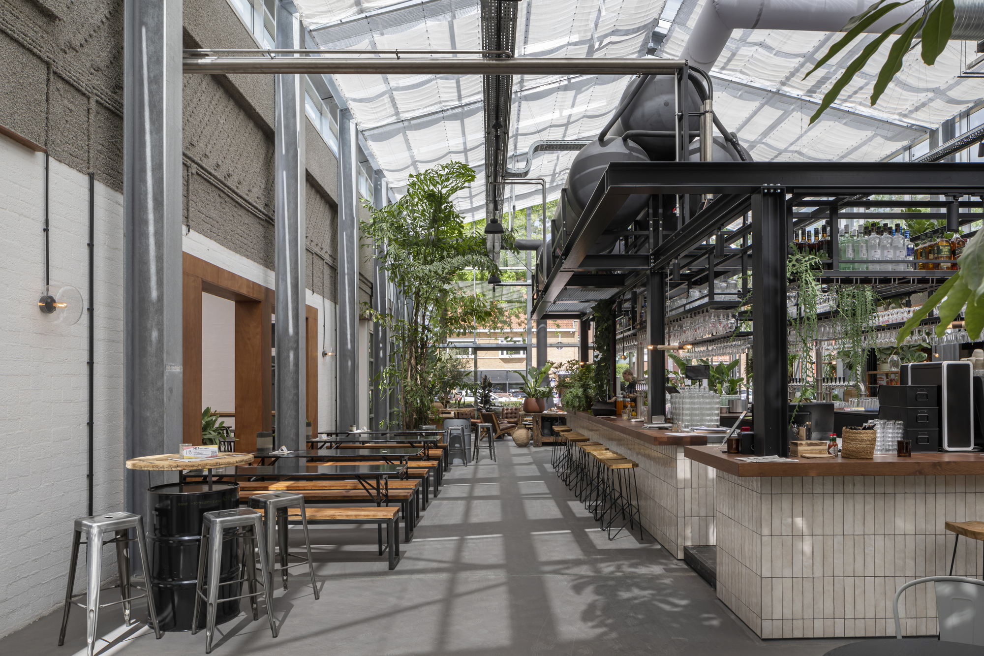 Interior dining hall with white tile bar, metal tables and benches beneath a translucent canopy roof
