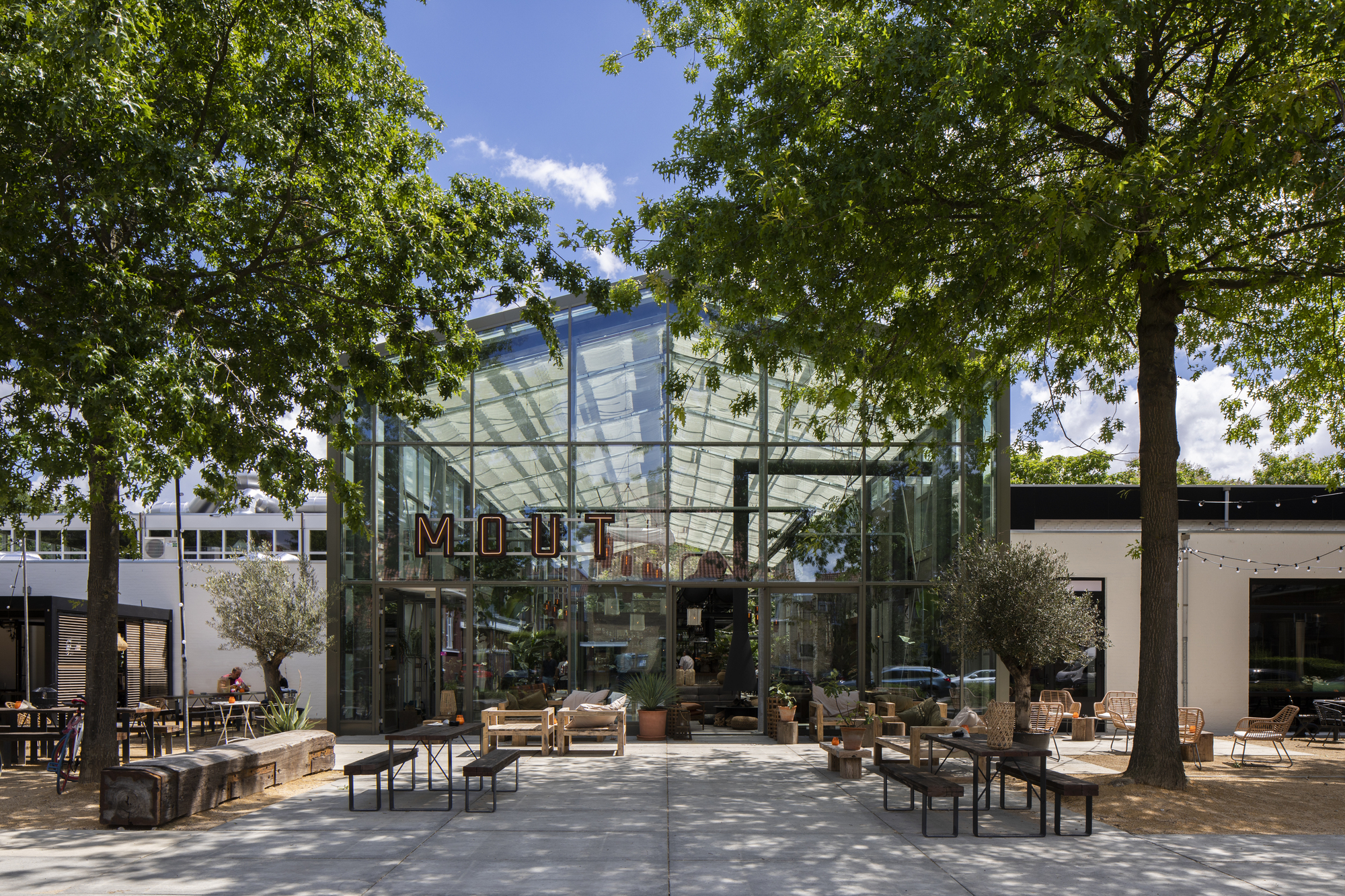 Glass pavilion entrance with steel frame and translucent canopy roof beneath mature trees on a sunny day