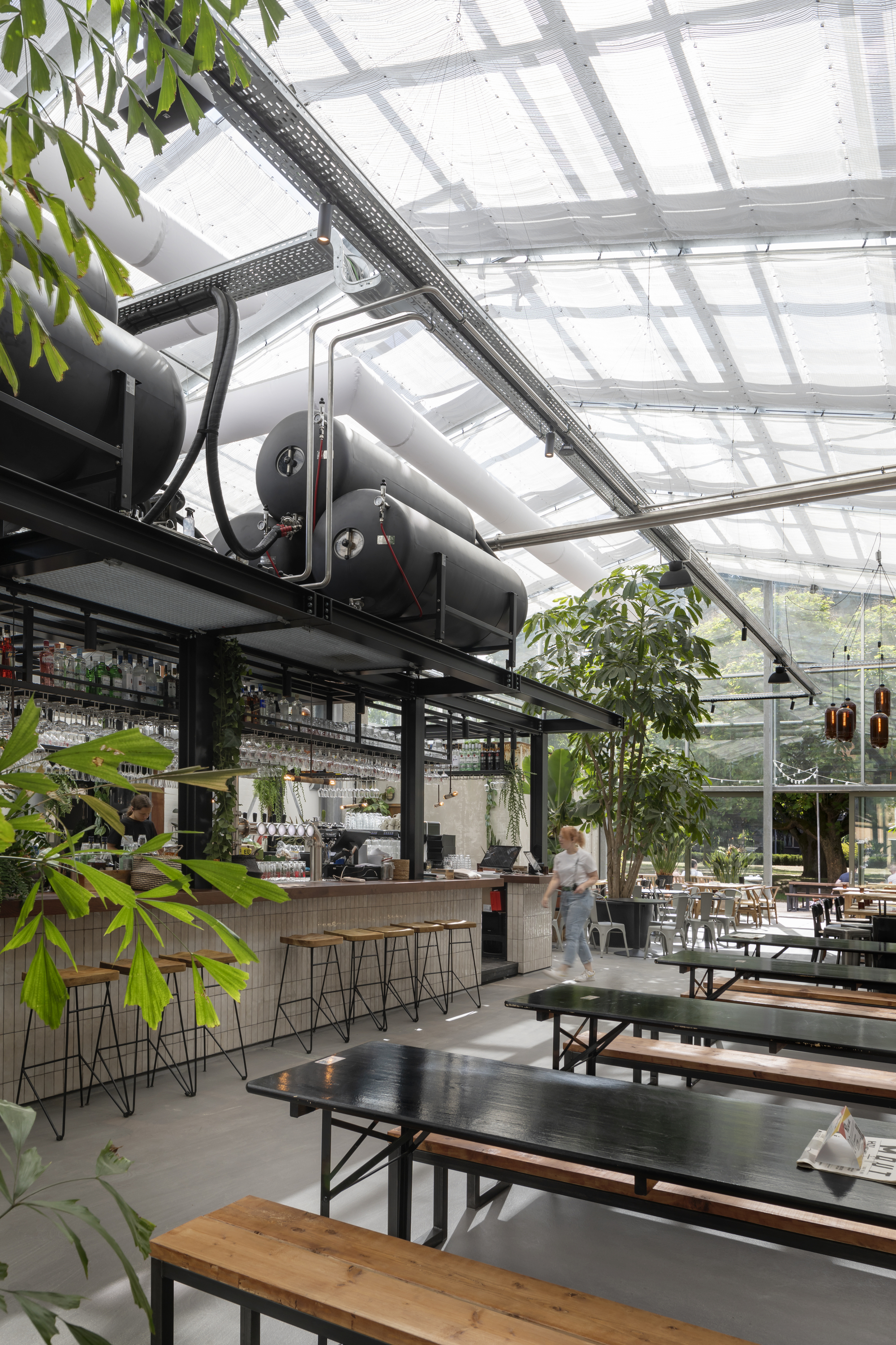 View of exposed black ductwork and steel frame supporting the translucent roof above dining tables and greenery