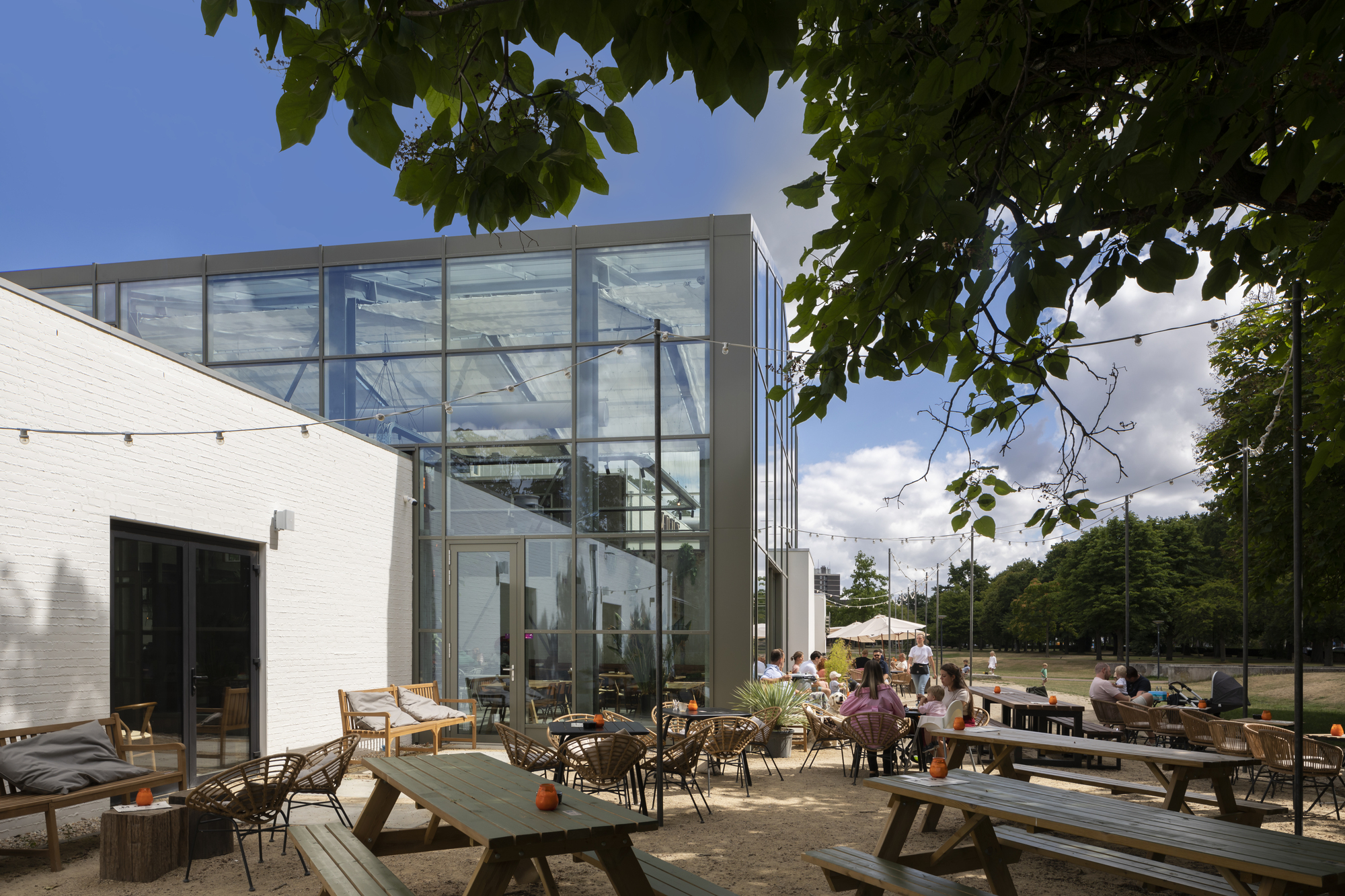 Two-story glass box addition connecting to white masonry volume with outdoor terrace seating under tree canopy