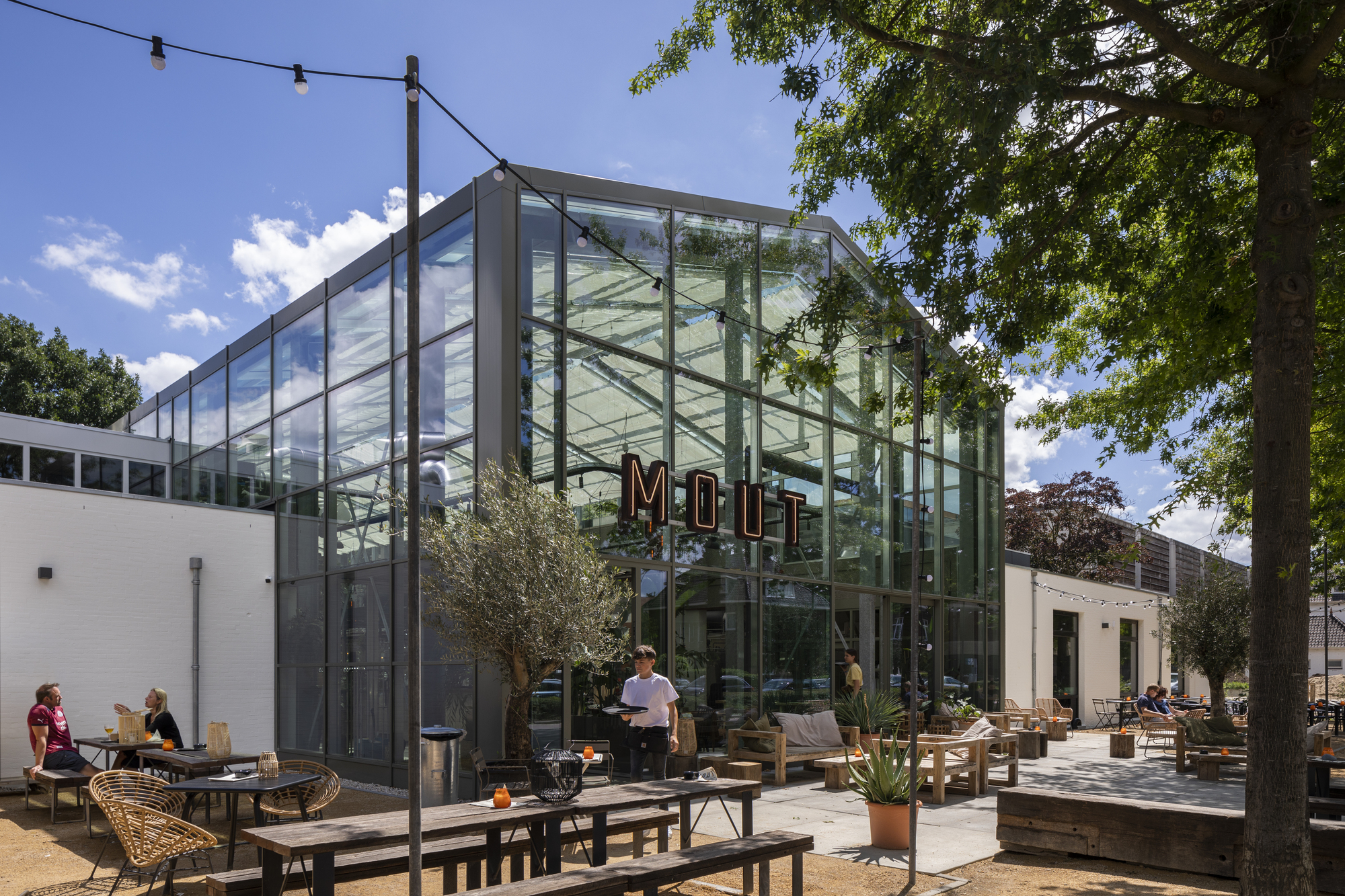 Glass-walled brewery pavilion with outdoor terrace seating under a blue sky