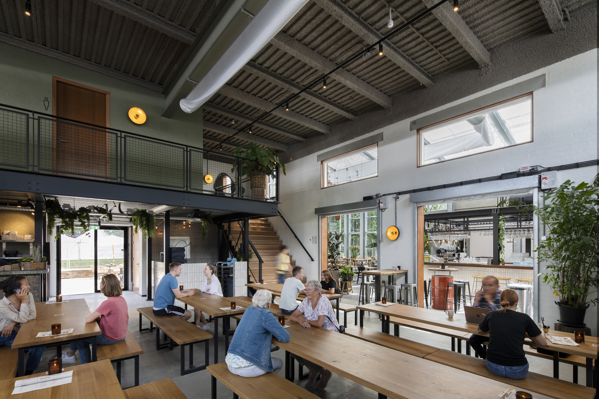 Double-height taproom with mezzanine level and patrons gathered at communal timber benches