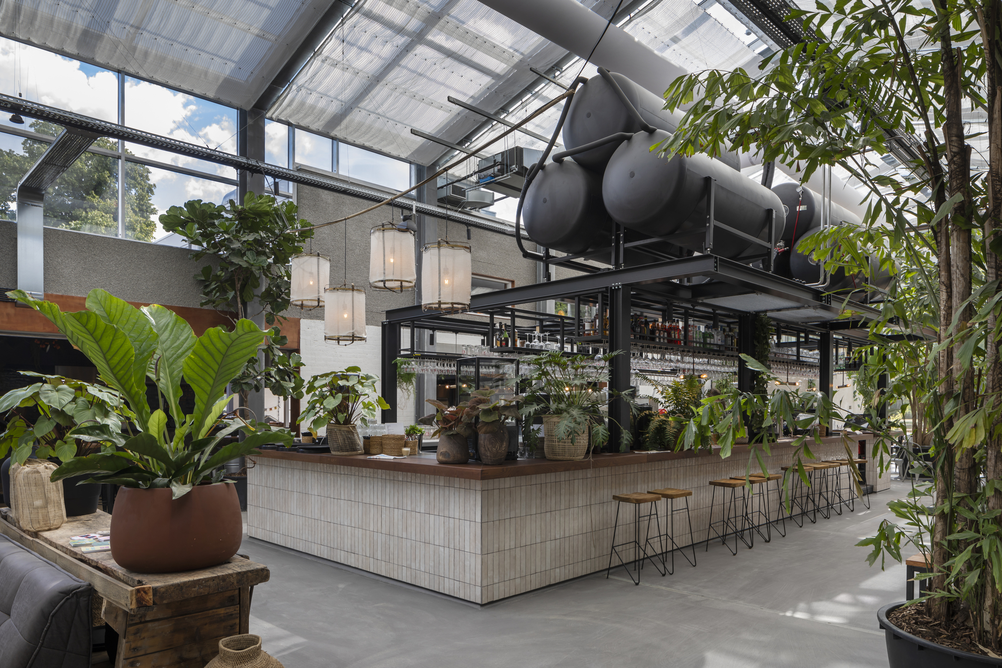 Tiled bar counter beneath exposed mechanical ductwork and skylights with potted plants throughout