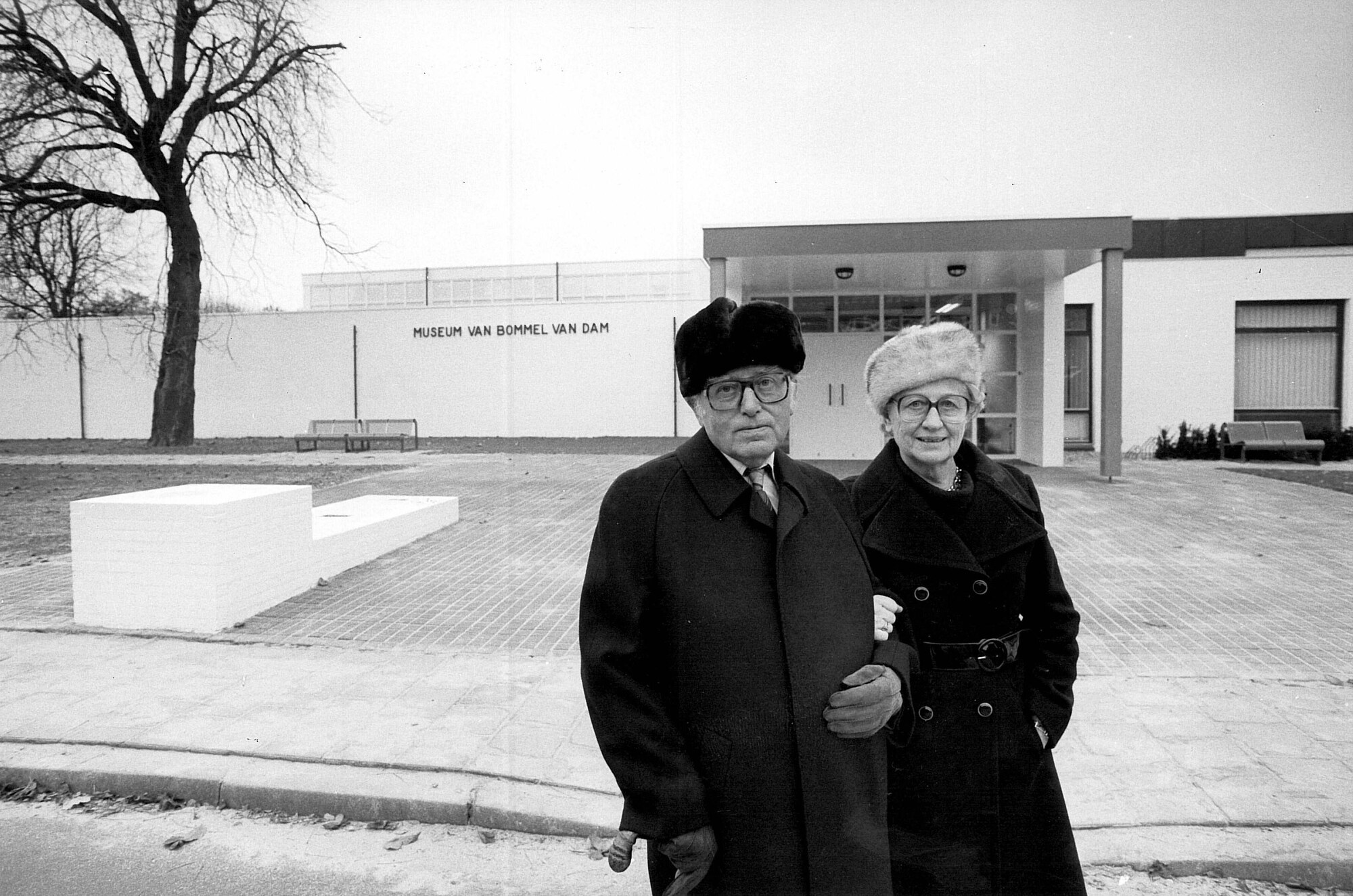 Exterior view of white-walled entrance pavilion with two visitors in winter coats beside a bare tree
