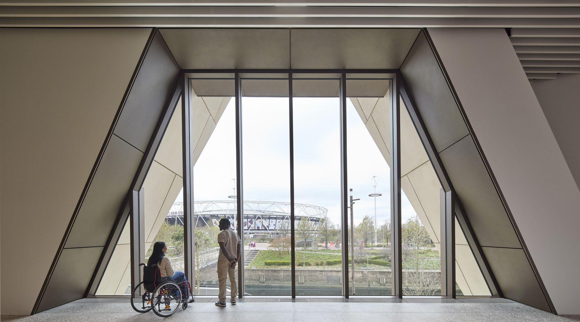 Interior gallery space with floor-to-ceiling triangular glazing overlooking the landscape and two visitors conversing