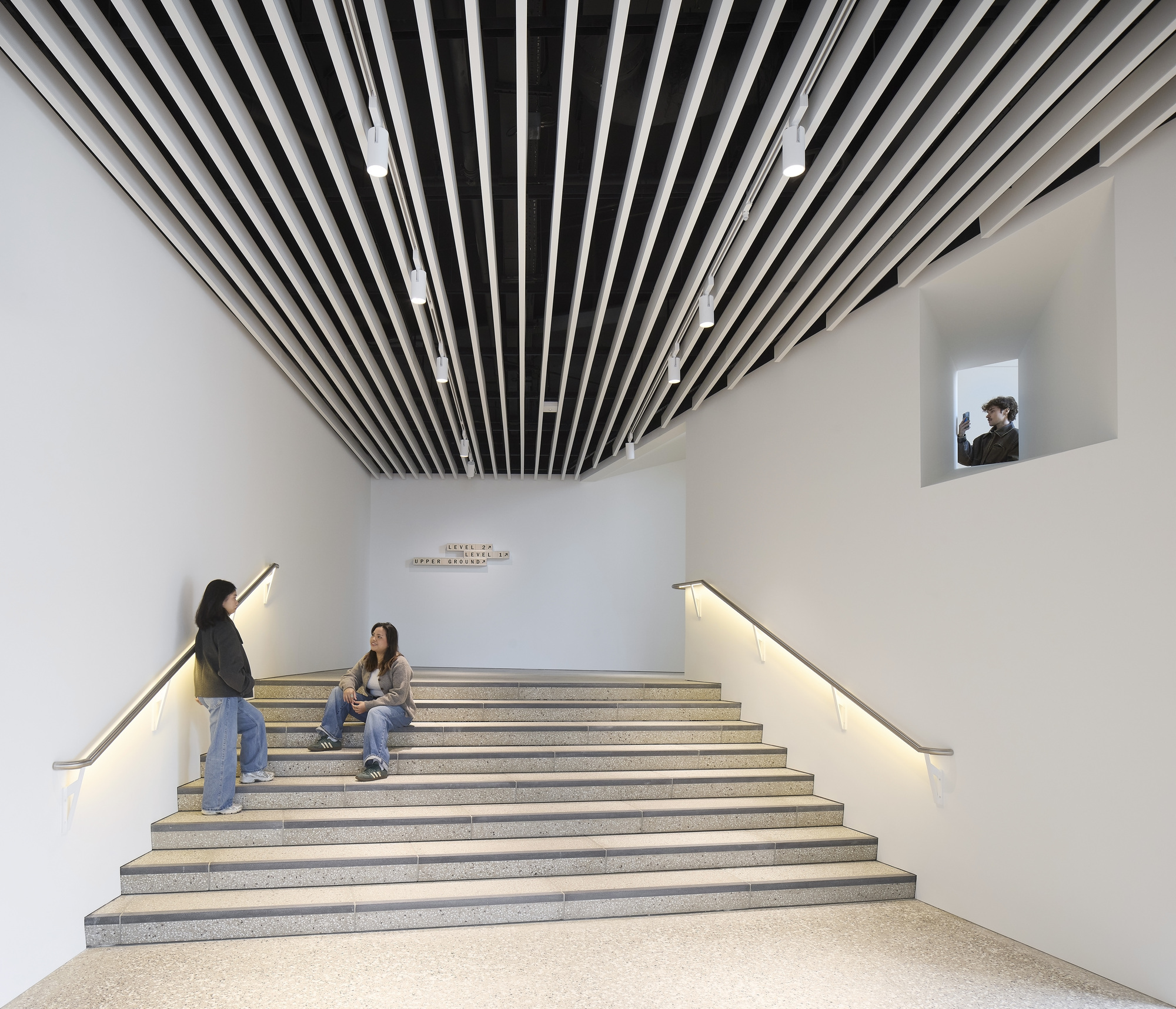 Wide staircase with illuminated handrails beneath radiating slatted ceiling baffles and visitors seated on the steps
