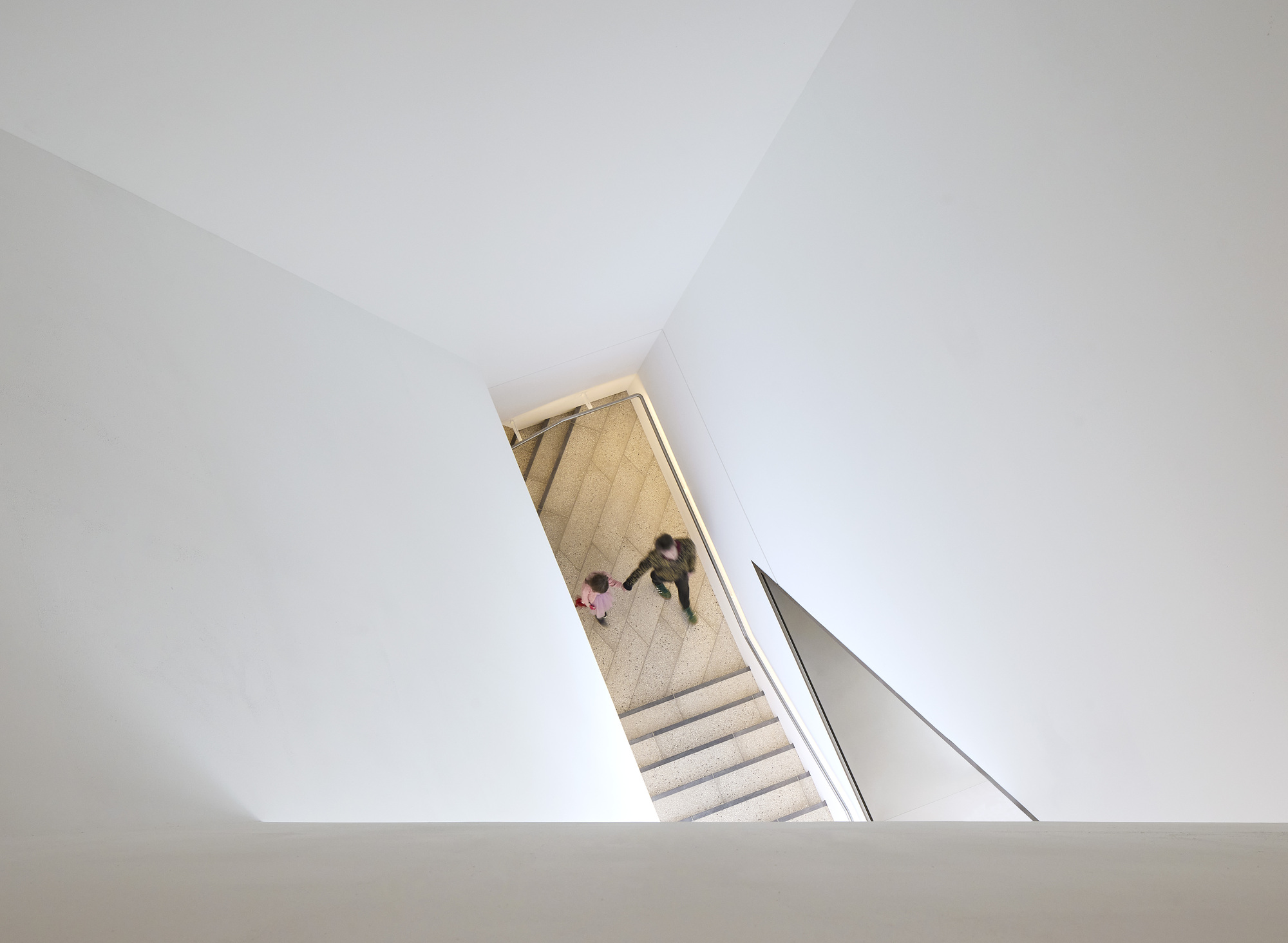 Overhead view of the white stairwell with figures descending the stone treads between angled walls