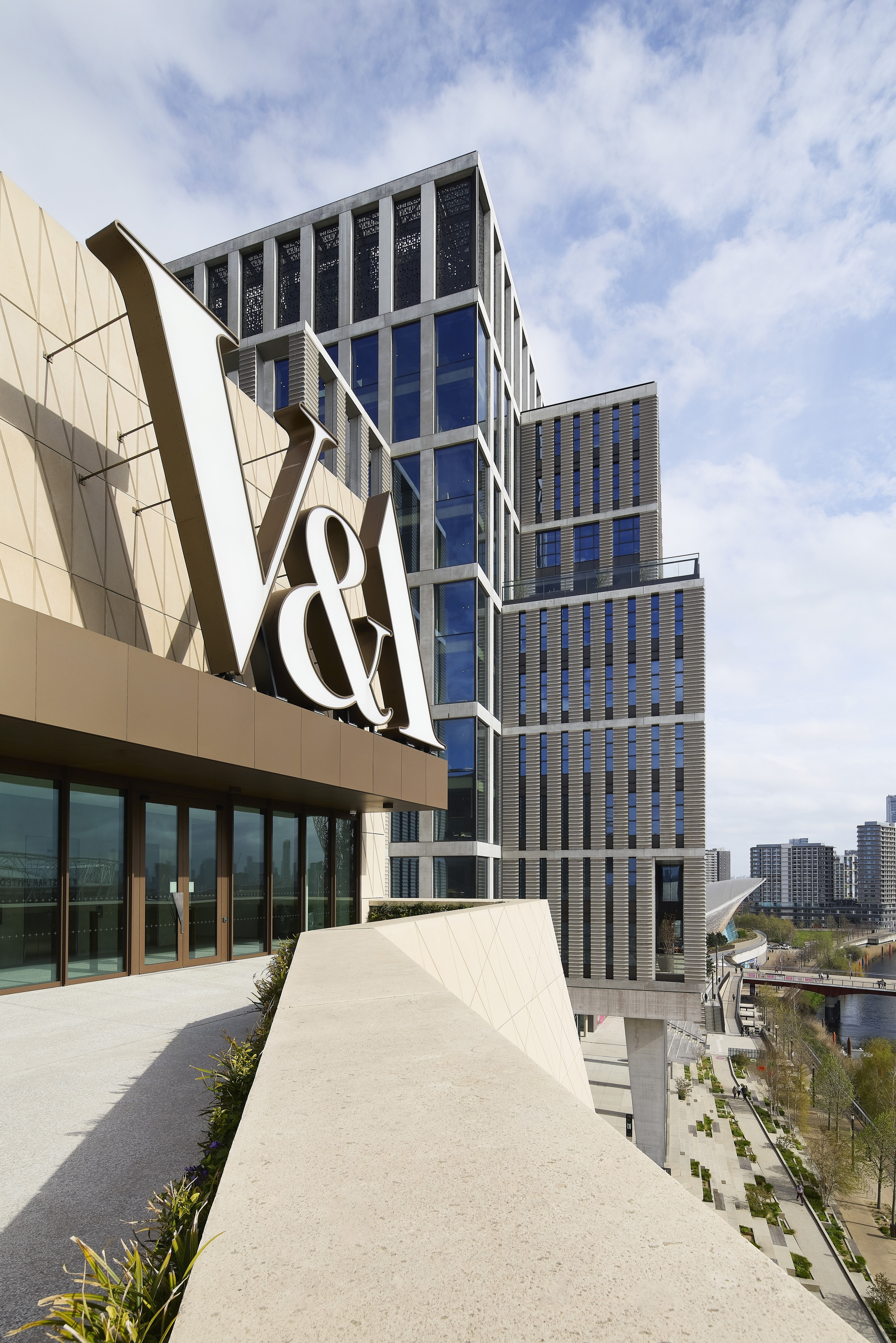 Rooftop terrace with geometric paving and angled facade forms rising beside a stepped glass tower under cloudy skies