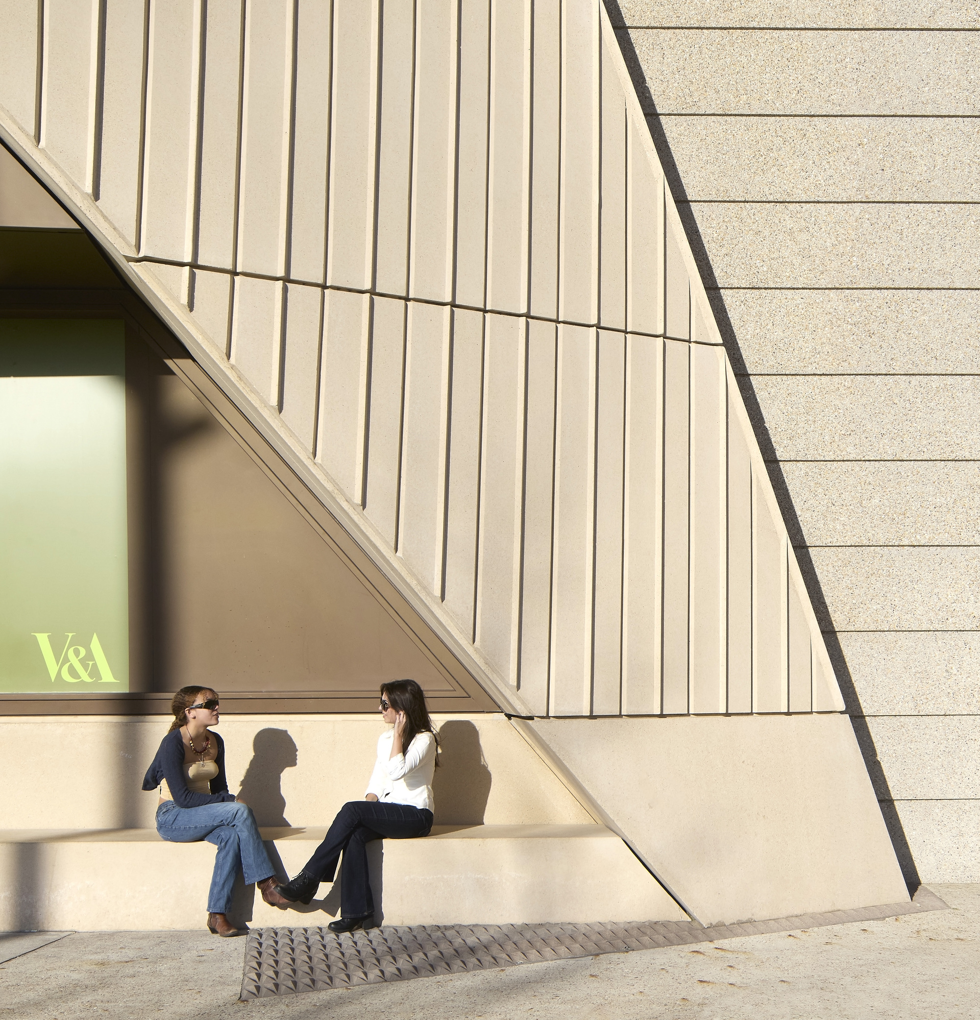 Exterior corner with vertical fluted panels and textured stone where two visitors sit in midday sun