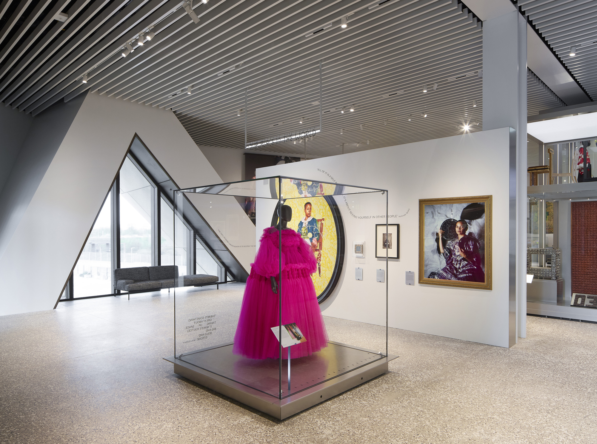 Gallery interior with slatted ceiling and magenta garment in glass vitrine beside angled glazing