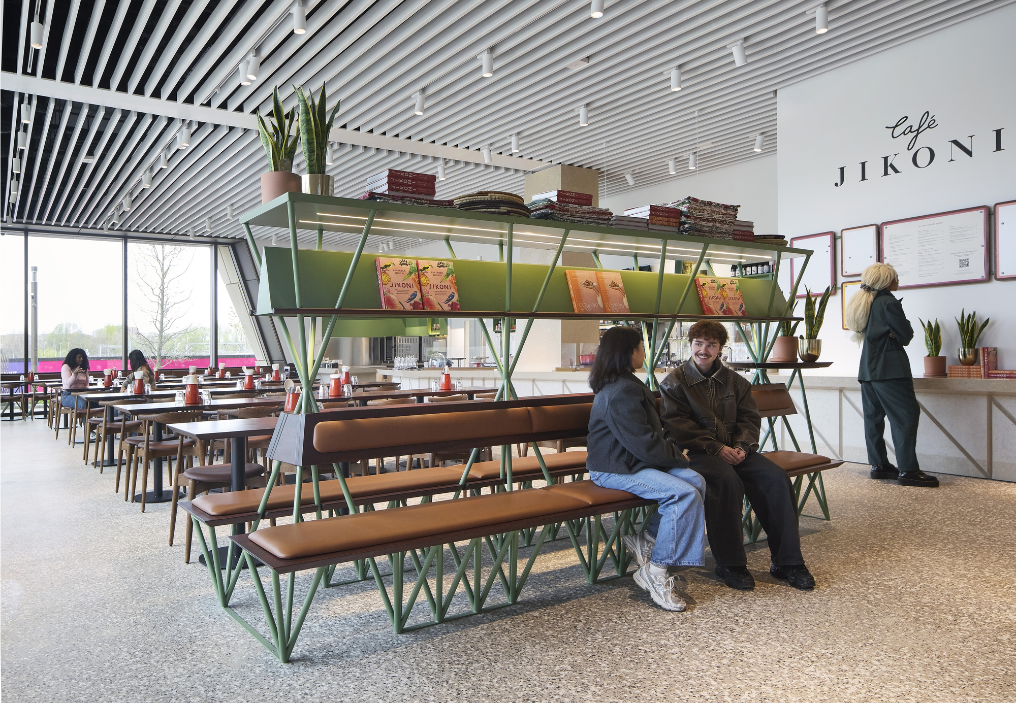 Cafe seating with green metal bench and shelving unit beneath a slatted ceiling with visitors nearby