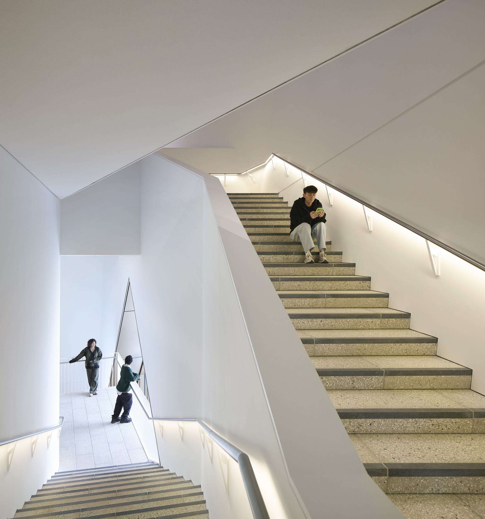 Multi-level stairwell with terrazzo treads and integrated handrail lighting as visitors descend and pause