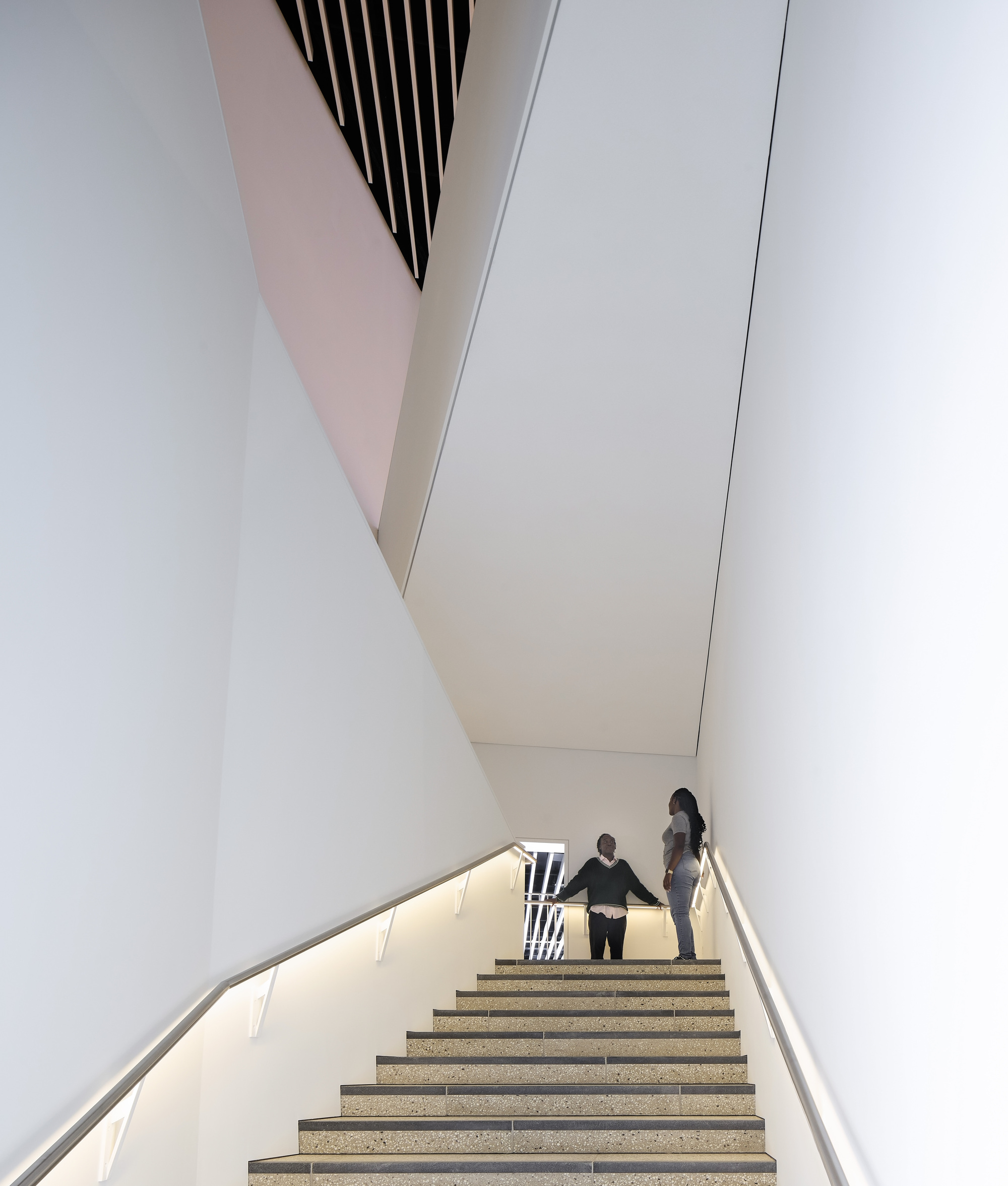 View up the angular stairwell with pink-tinted walls and two figures ascending toward daylight