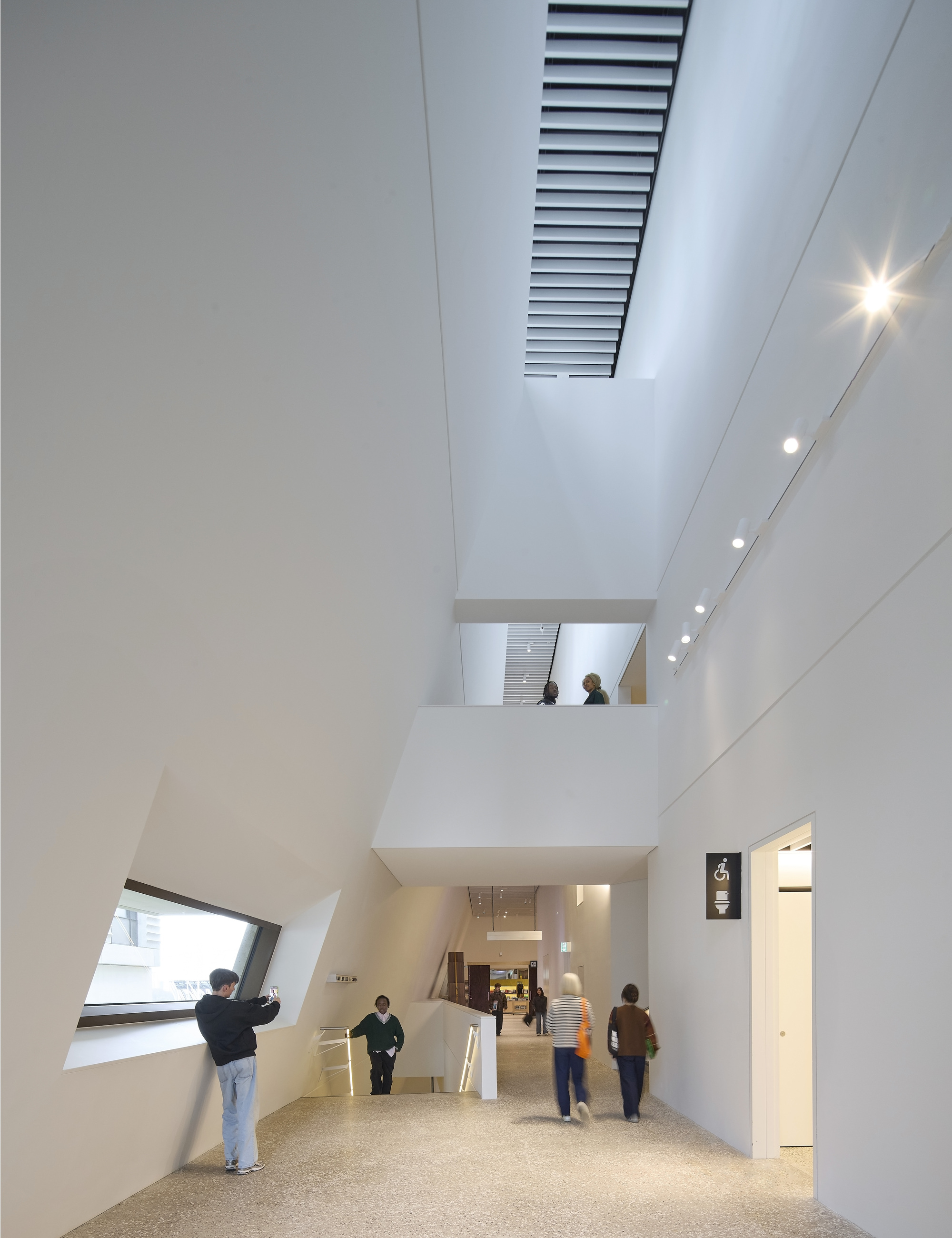 Double-height atrium with angled window, internal balcony and visitors moving through the terrazzo-floored circulation space