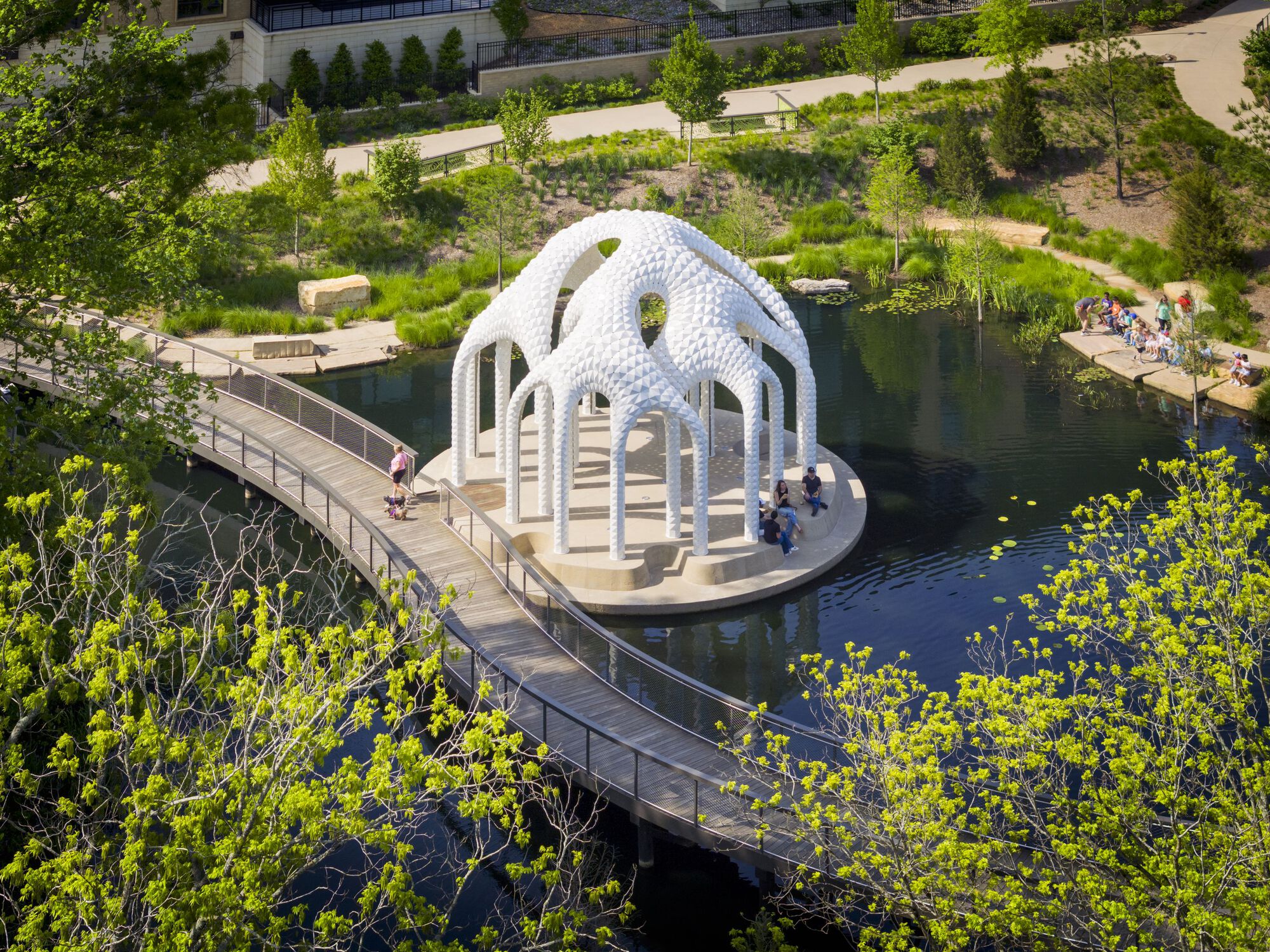 Aerial view of white pavilion with arched columns on circular platform in pond accessed by timber boardwalk