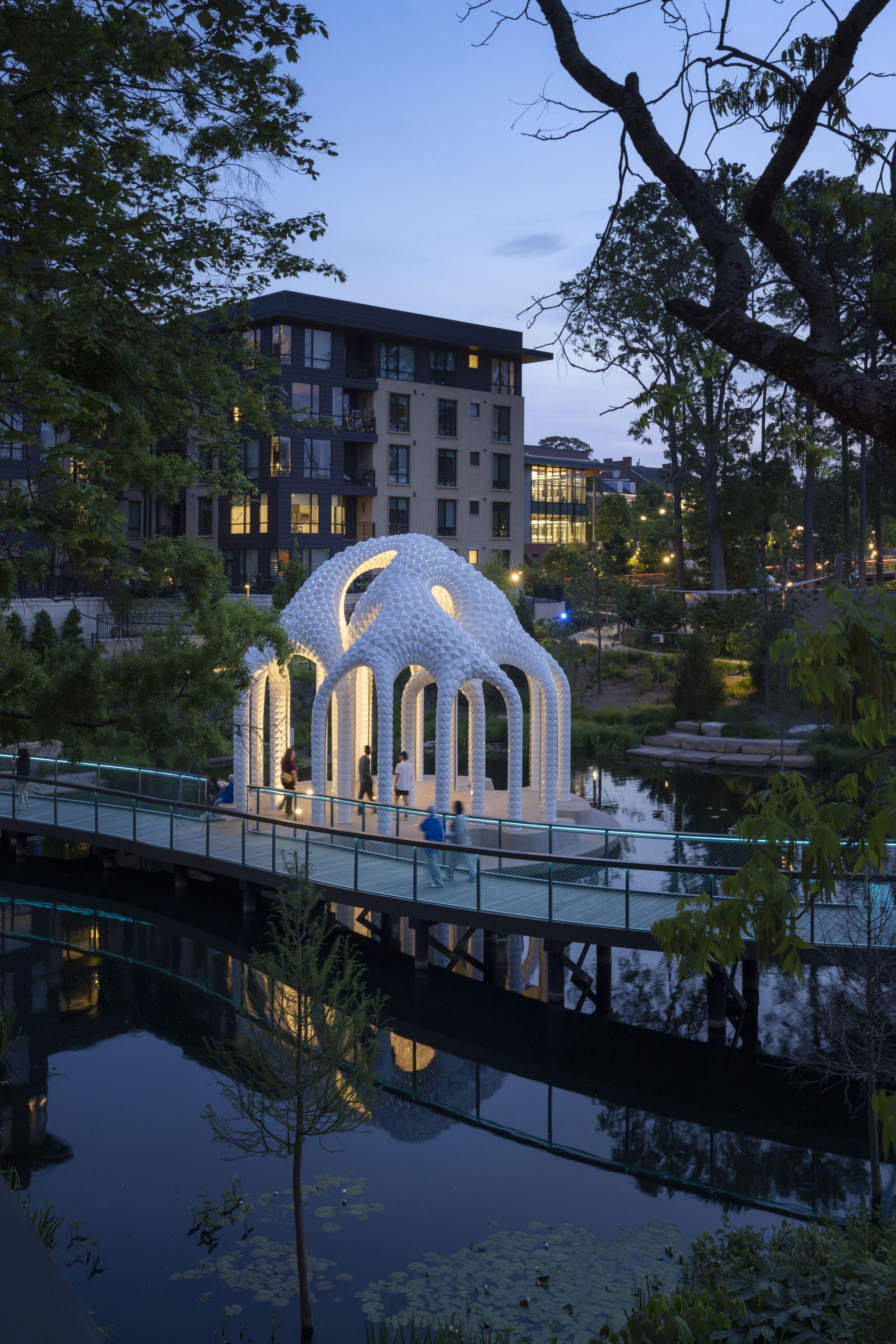Illuminated pavilion reflected in still pond water at dusk with residential buildings beyond