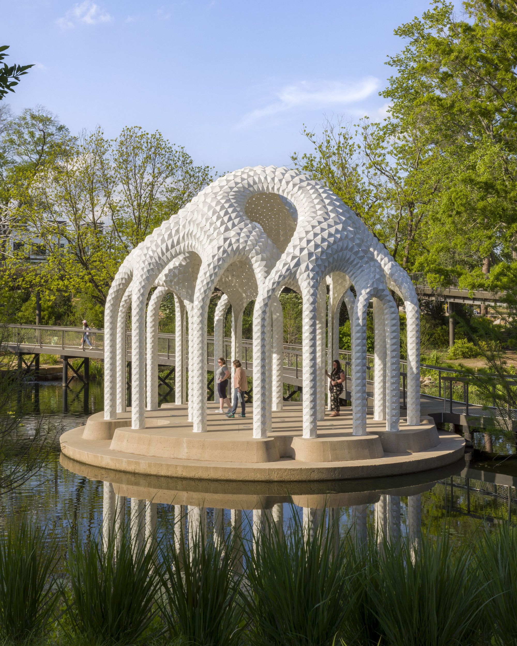 Pavilion with perforated white columns and curving roof on a circular platform over water surrounded by trees