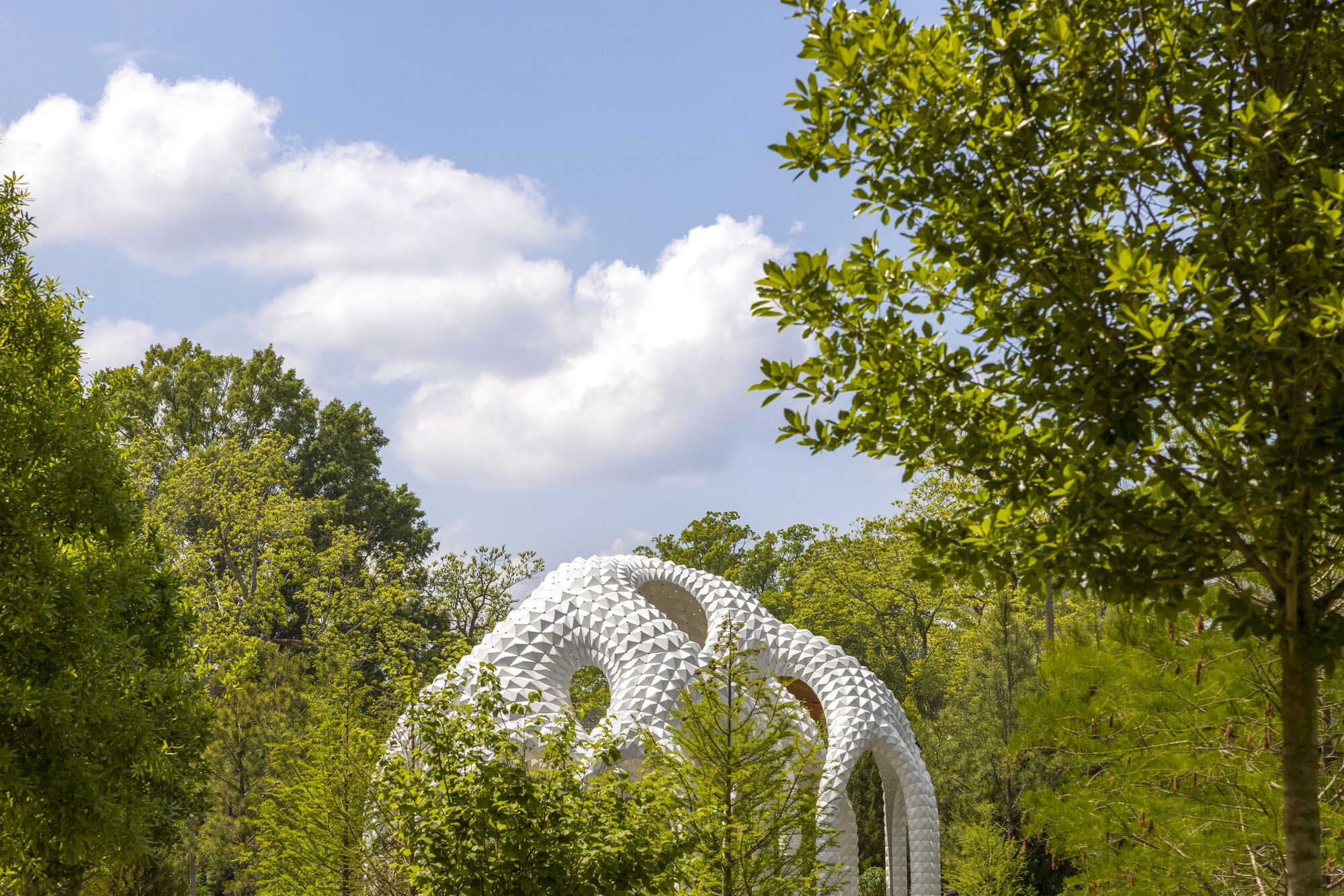 White sculptural loops rising above tree canopy on a partly cloudy summer day