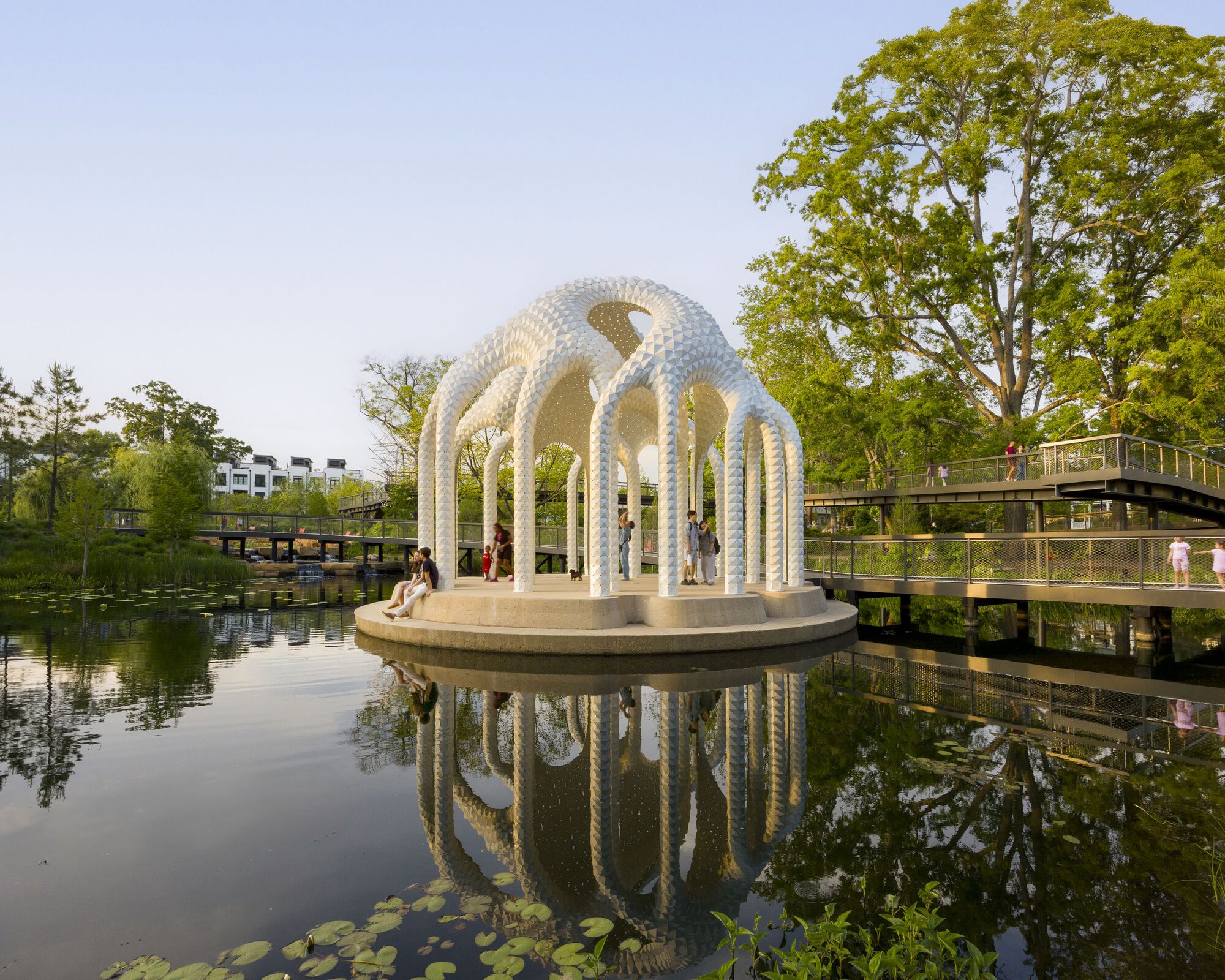 White latticed pavilion with arched openings on a circular platform reflected in a calm pond surrounded by trees