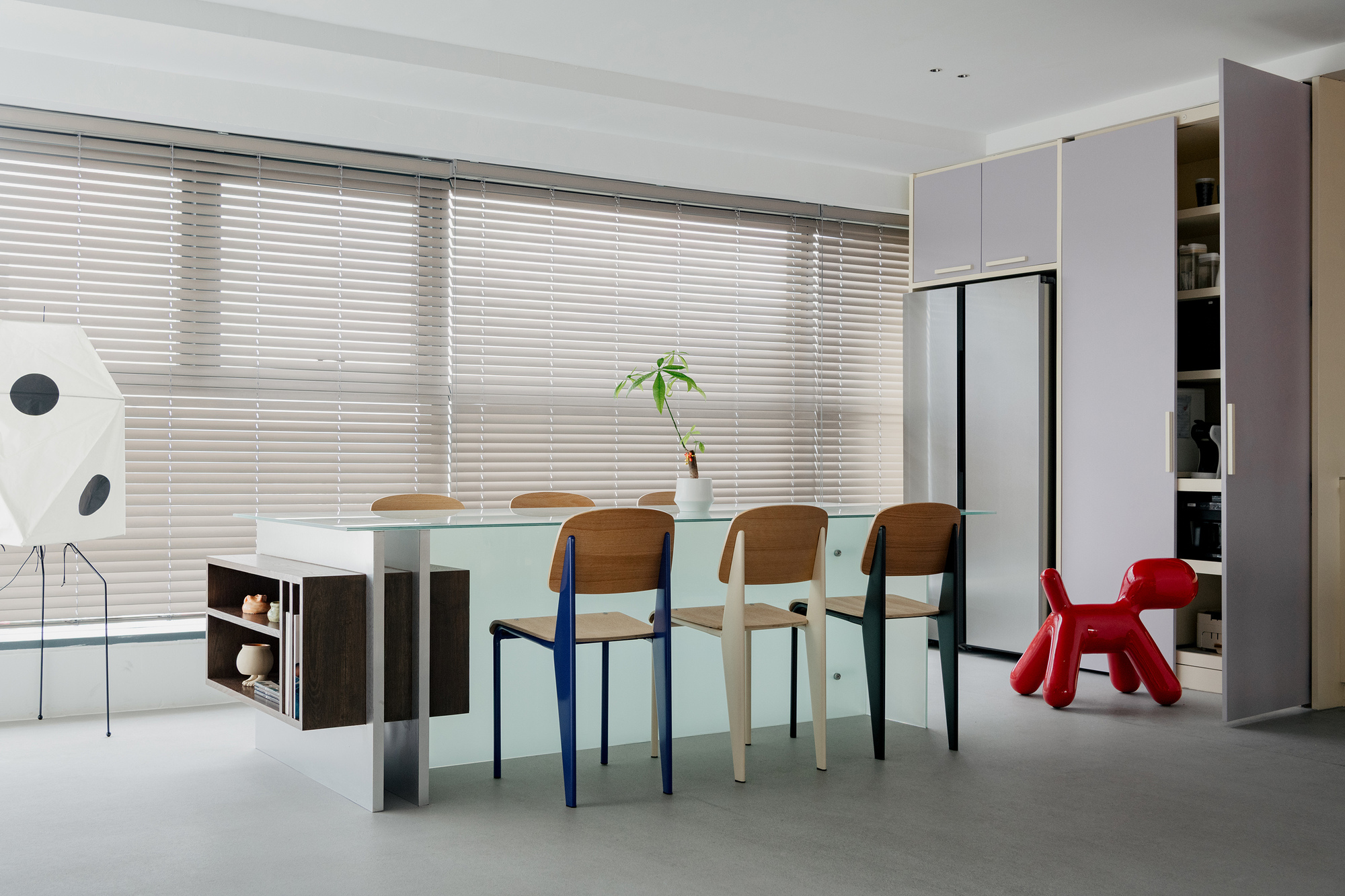 Dining area with frosted glass table, mismatched chairs, and horizontal blinds filtering daylight from floor-to-ceiling windows