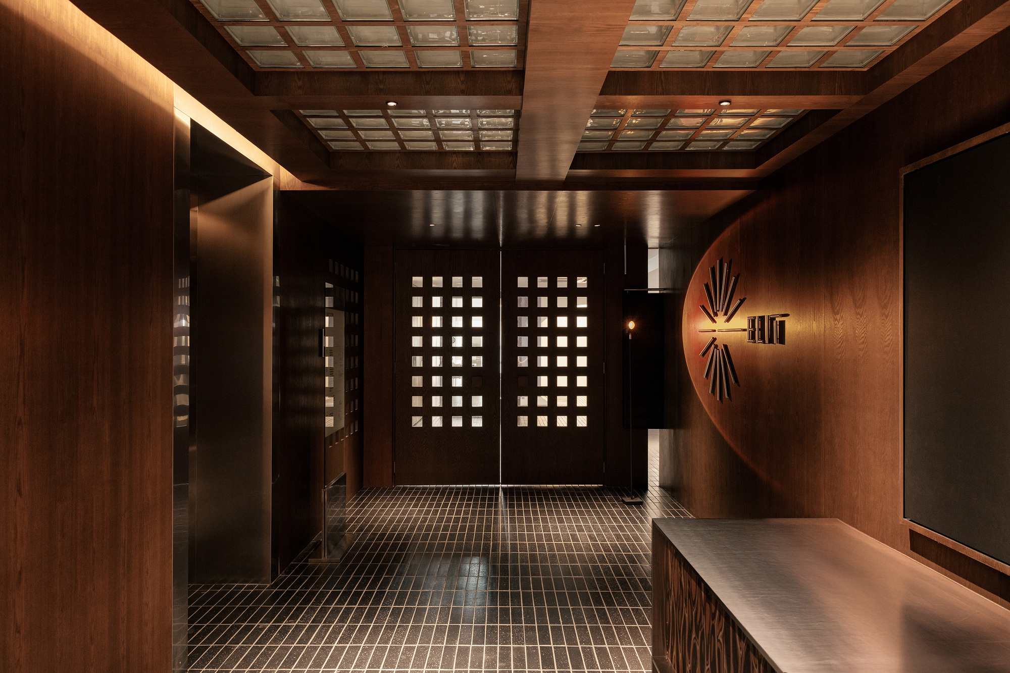 Corridor with perforated timber doors and gridded ceiling panels framed by dark mosaic floor tiles