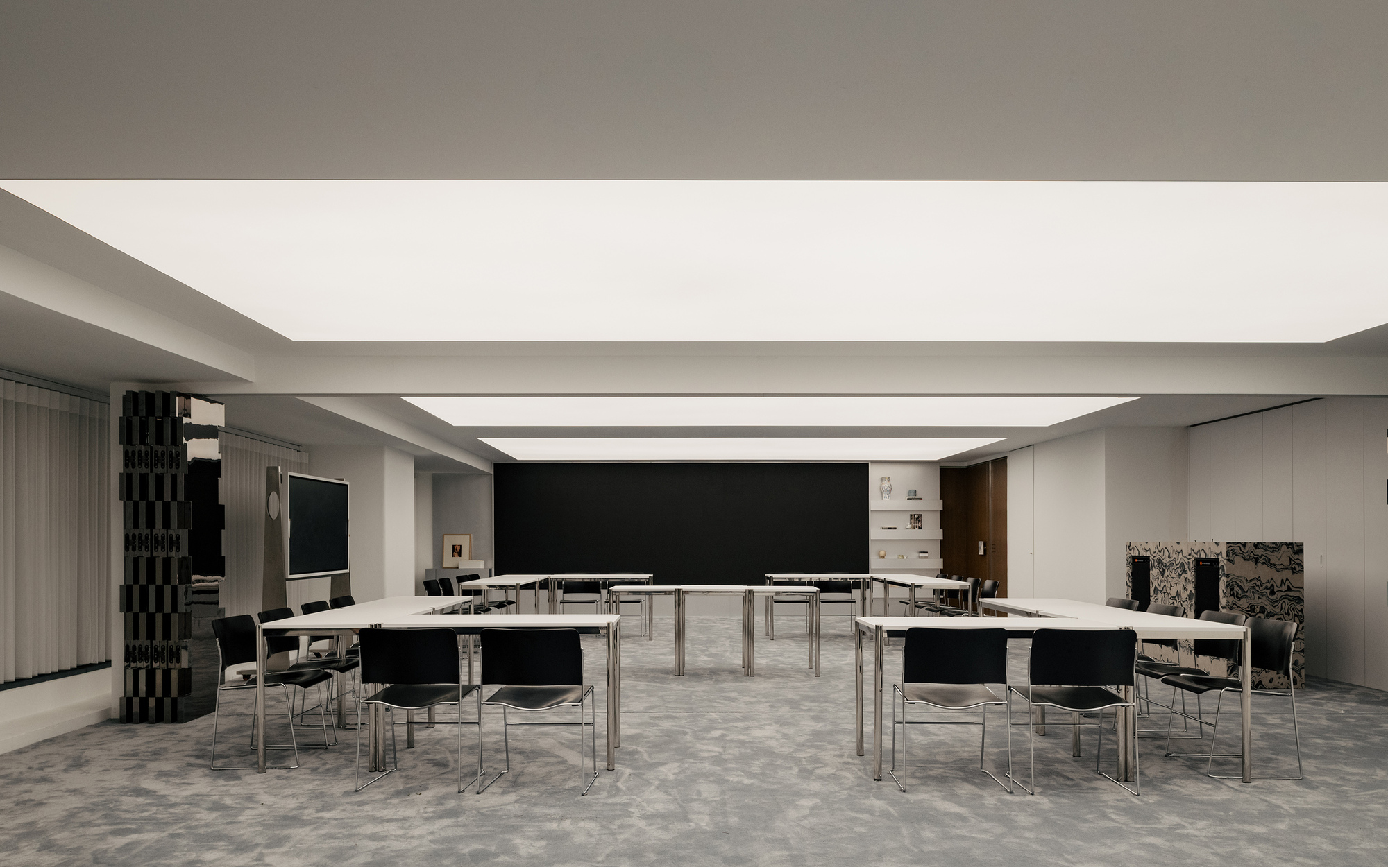 Meeting space arranged with white tables in clusters beneath linear ceiling skylights and patterned grey carpet