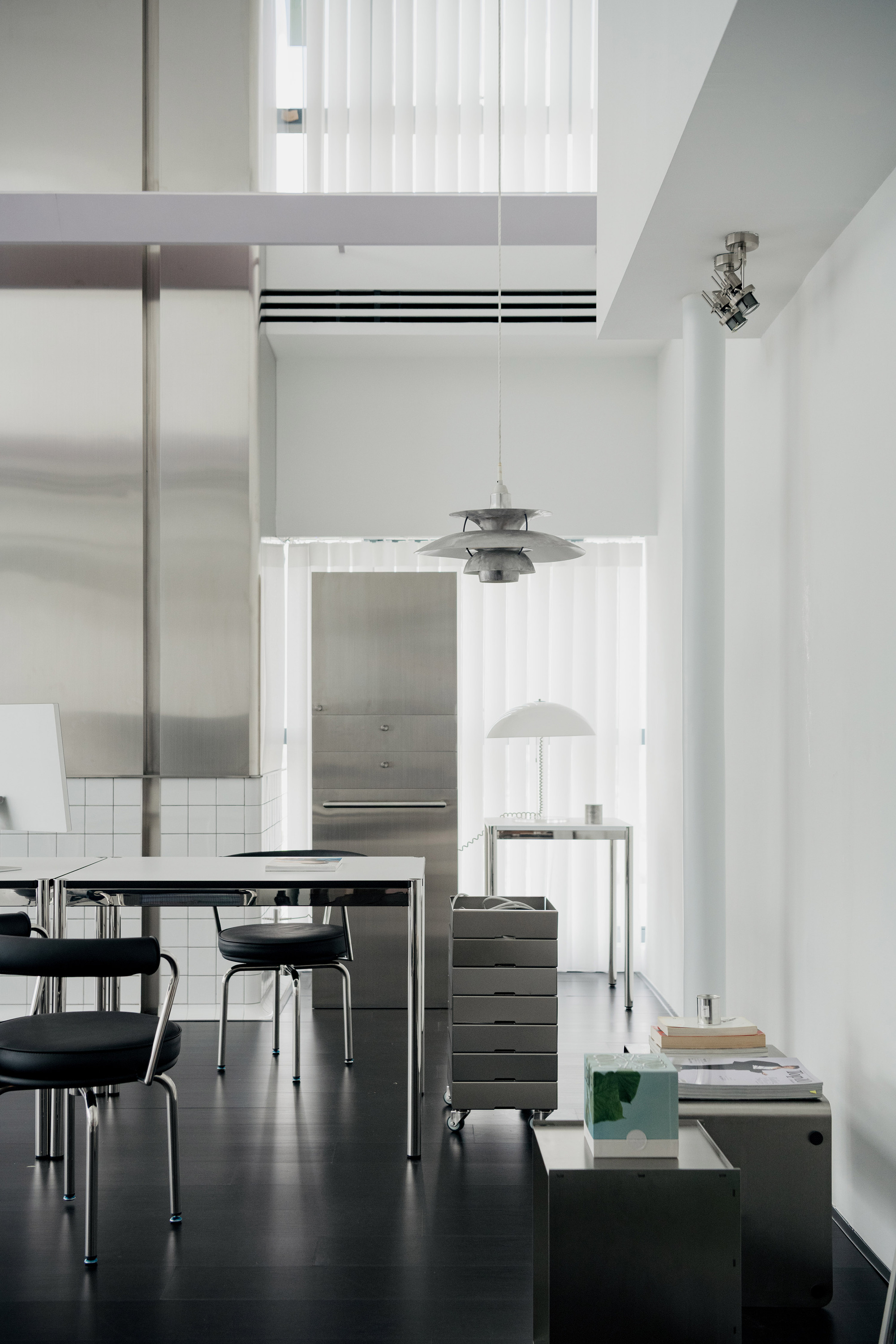 Double-height interior with dark flooring, steel furniture, pendant lamps and white vertical panel partitions