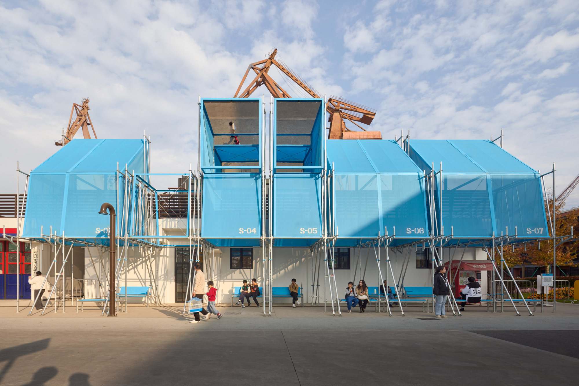 Stacked blue shipping containers with open doors and visitors seated on benches beneath
