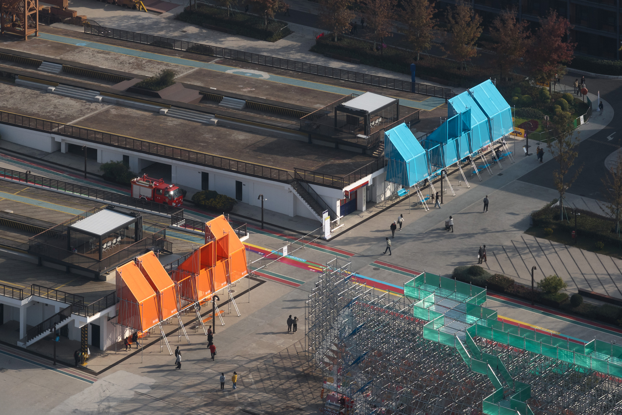 Aerial view of three colored pavilions on a plaza with pedestrians and nearby rail tracks