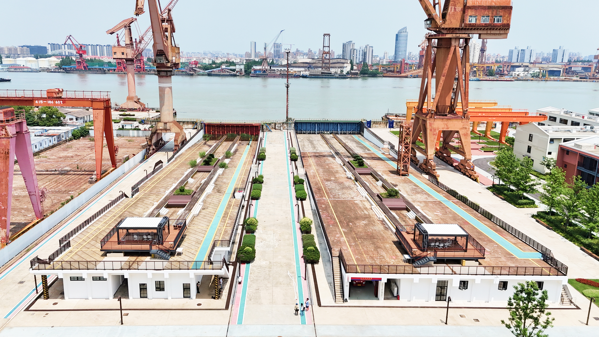 Elevated view of linear industrial site with warehouses, planted rows, and harbor cranes beyond