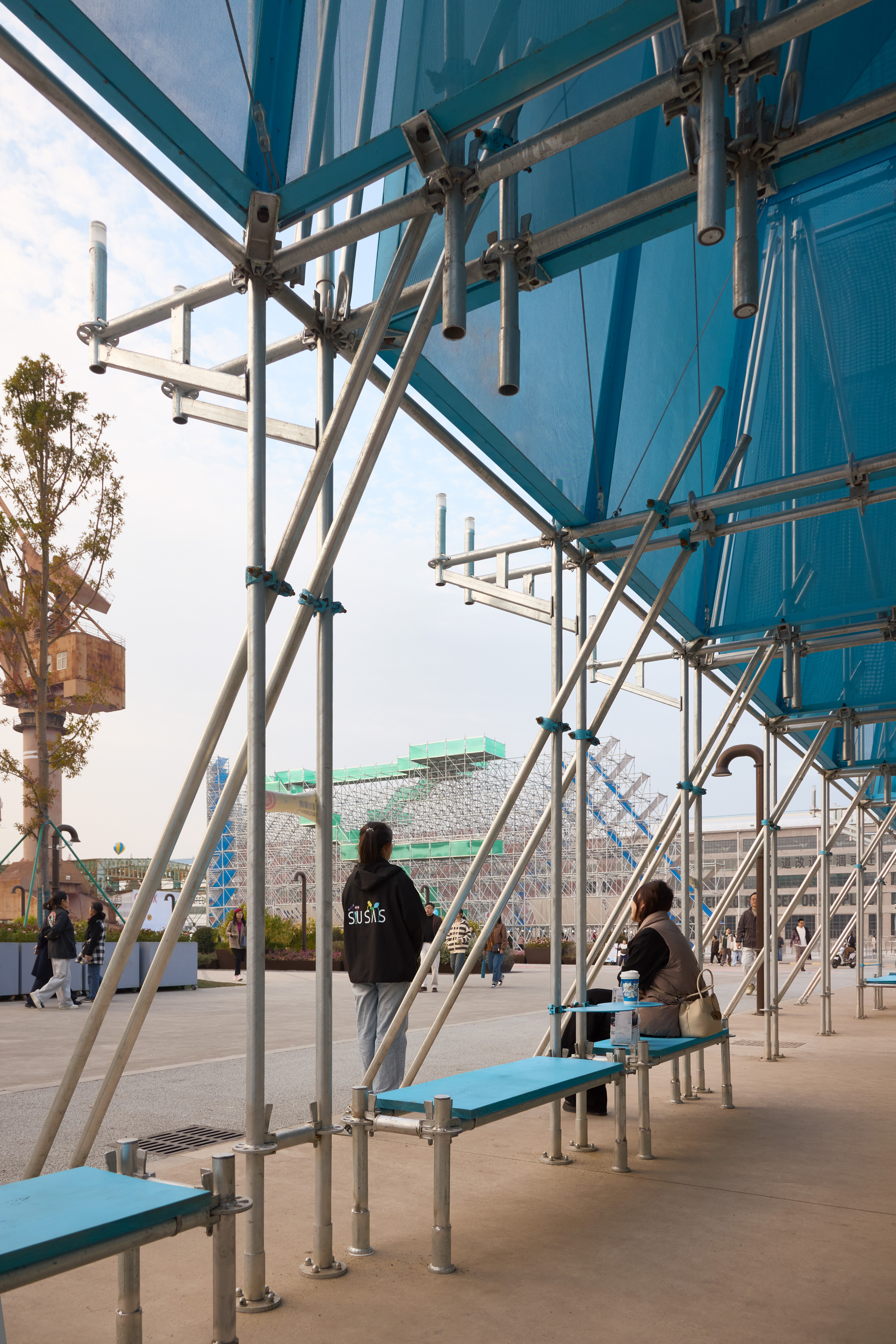 Scaffolding benches beneath a blue polycarbonate canopy where visitors sit in afternoon light