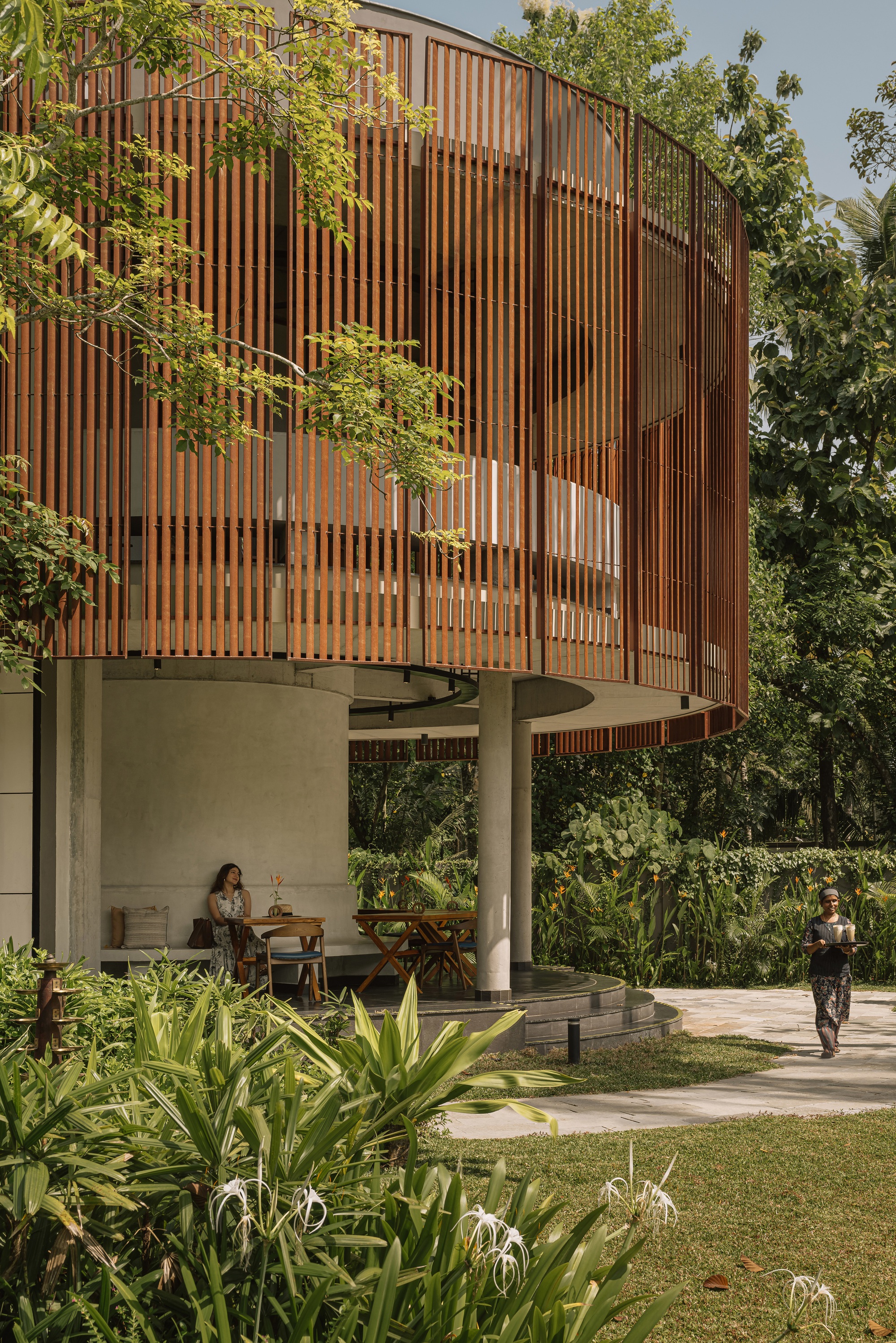 Elevated timber slat screen above concrete columns sheltering an open-air dining terrace with two figures walking
