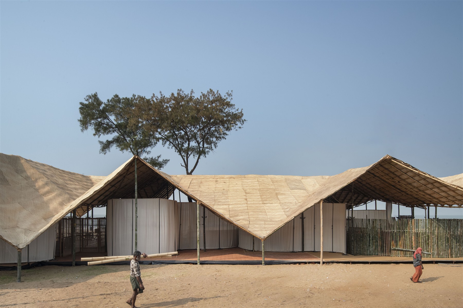 Woven bamboo roof structure with zigzag peaks rising above a sandy courtyard with children playing