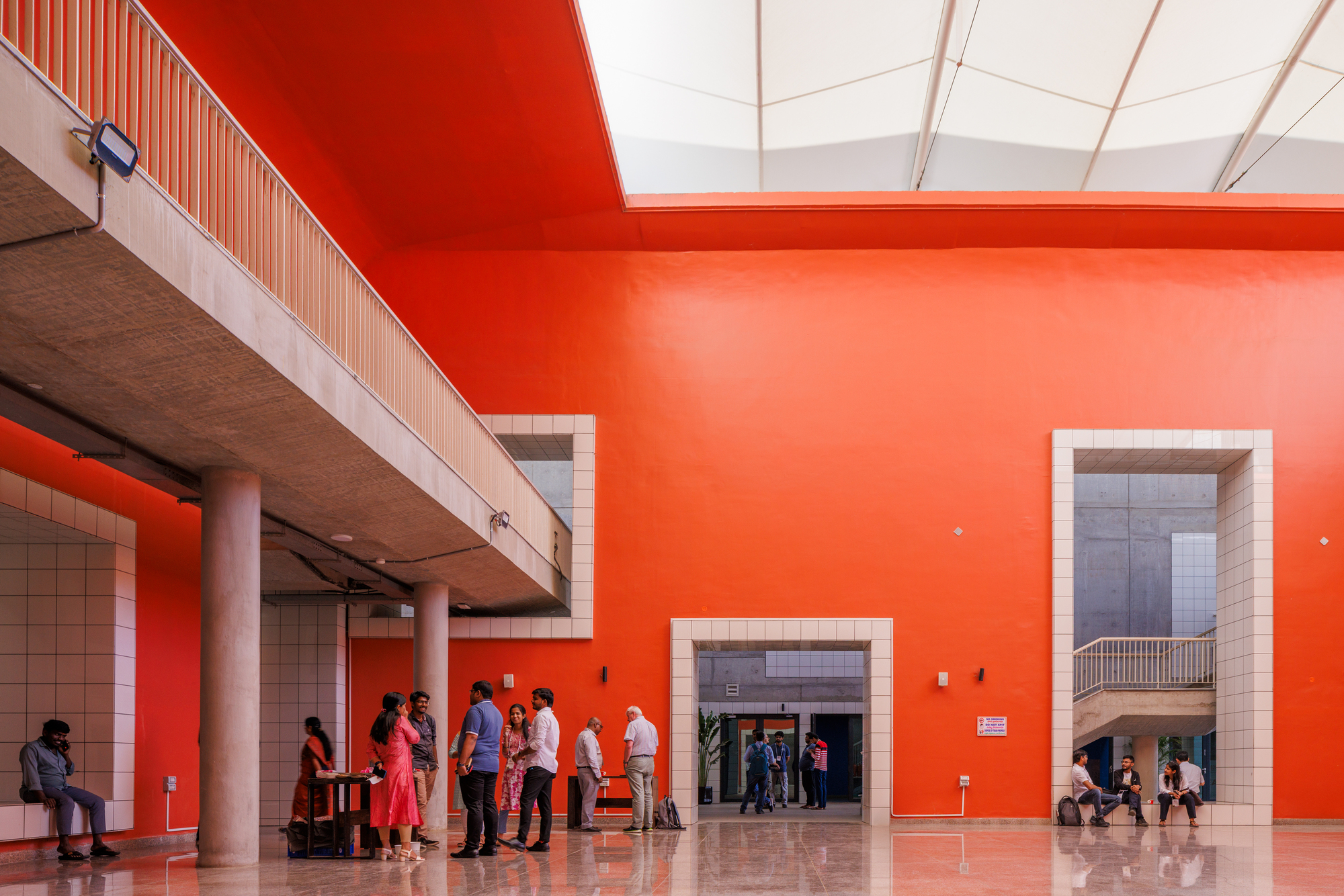 Interior hall with red-orange painted wall, white-framed openings, and groups of people conversing beneath skylights