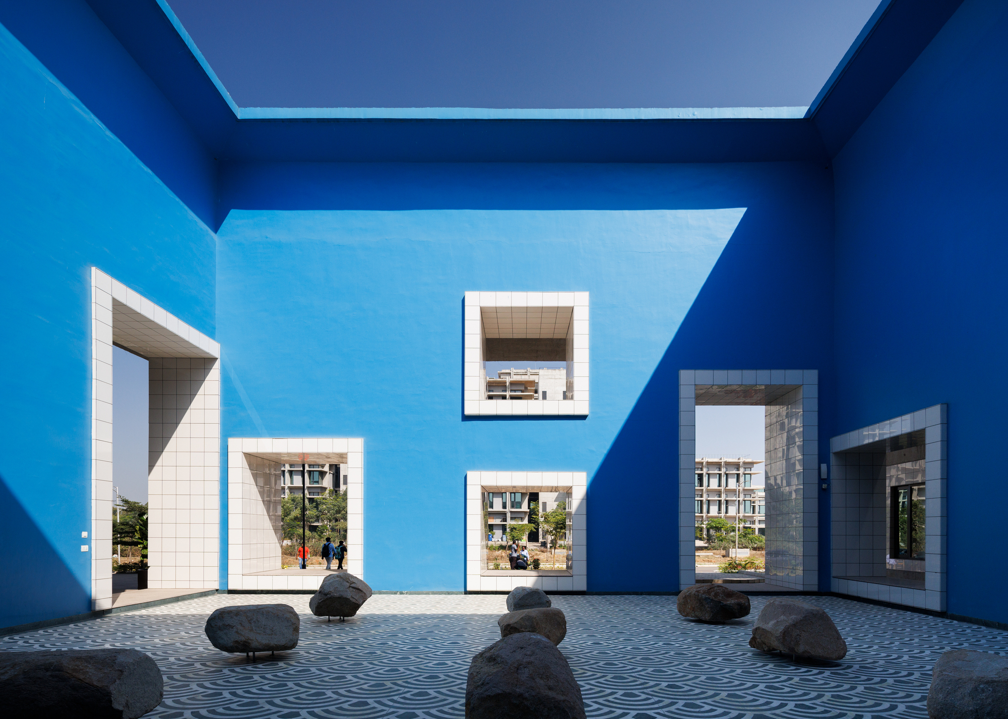 Blue-painted courtyard with white-framed windows, scattered rocks on tiled paving, and views through to planted greenery