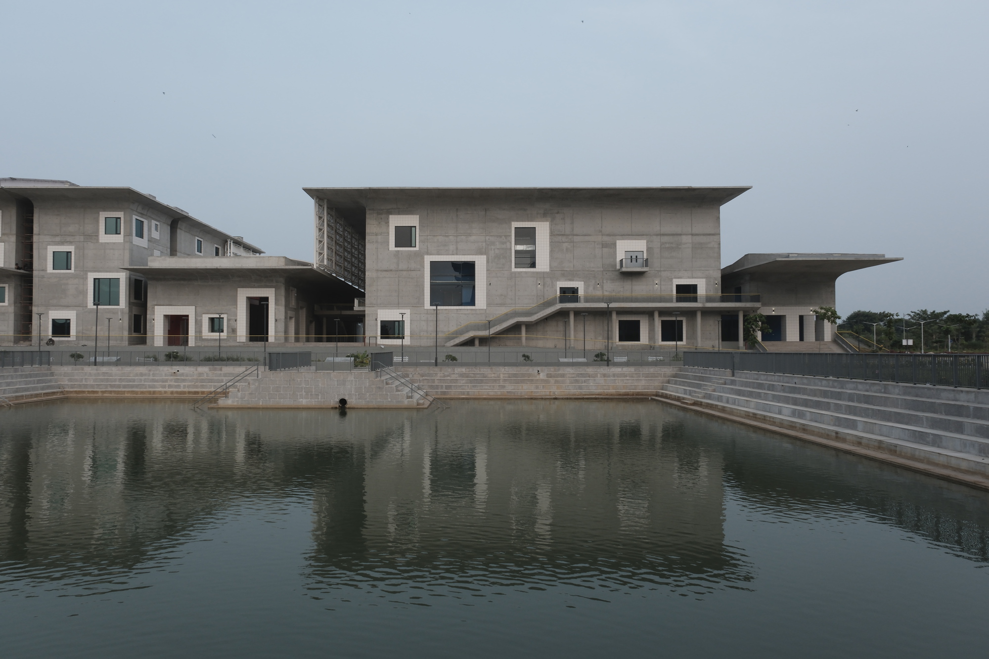 Concrete facade with scattered square windows reflected in a still pond under overcast sky