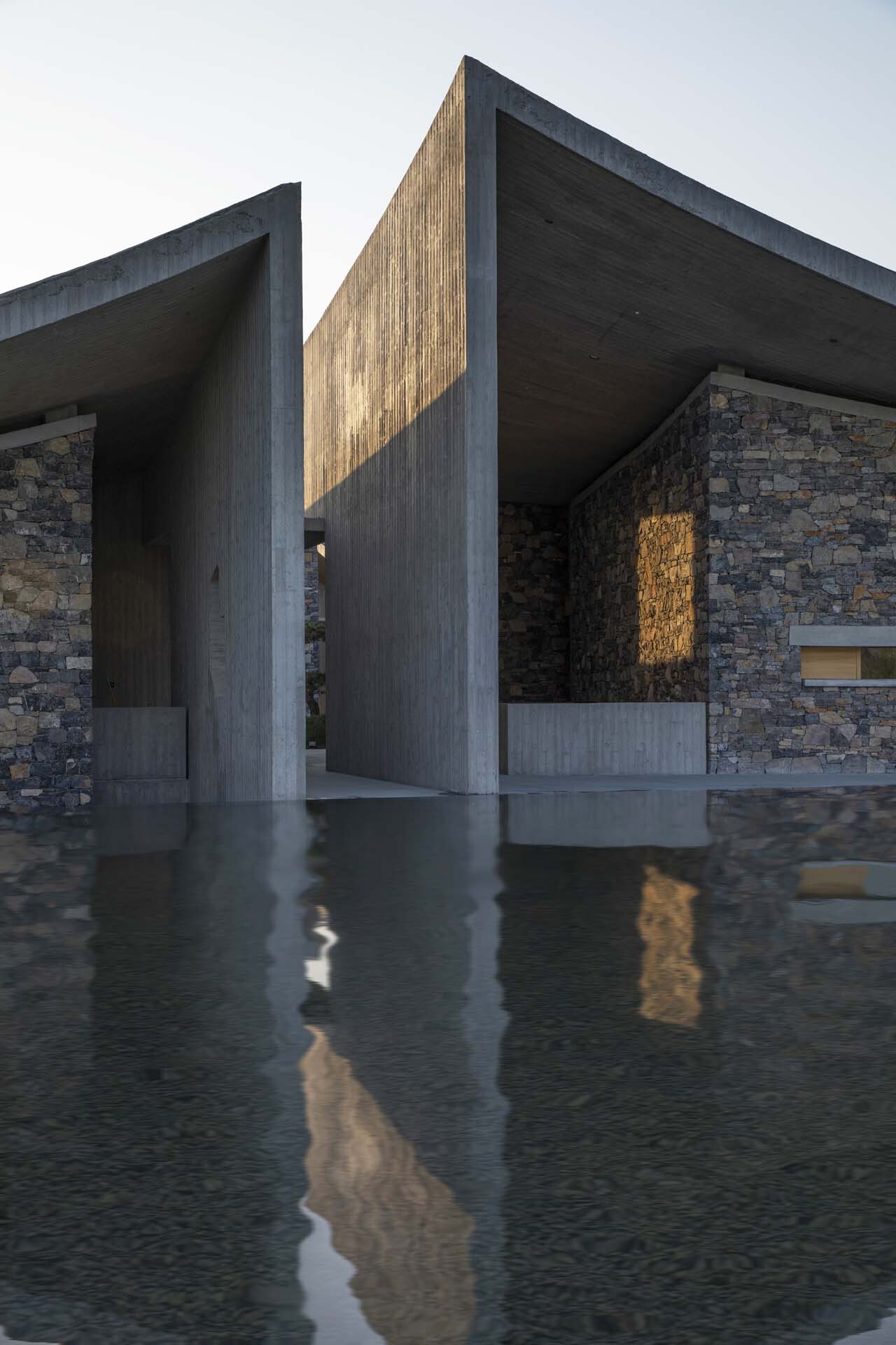 Covered entry passage with board-formed concrete columns and shallow reflecting pool catching late afternoon light