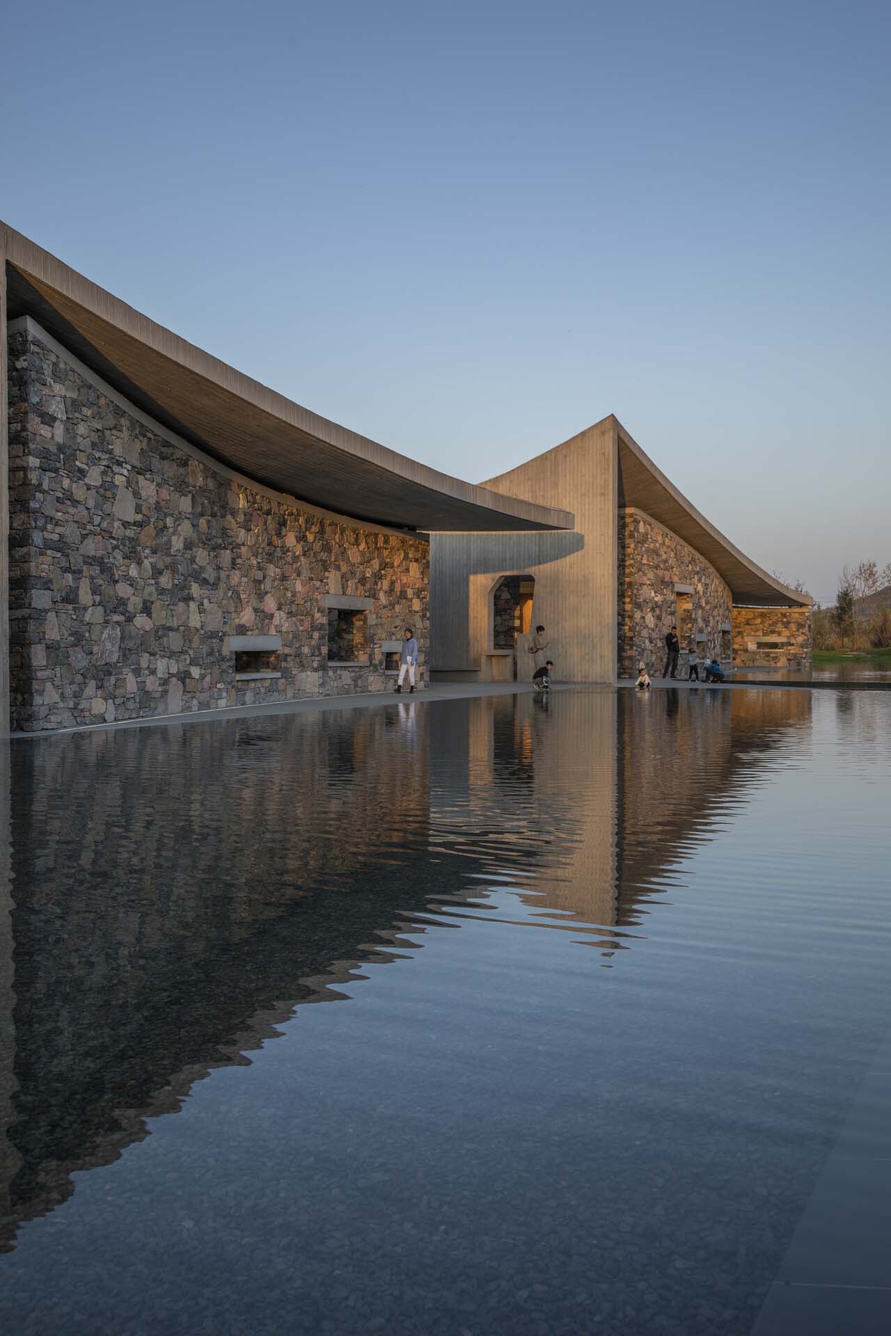 Stone masonry facade reflected in a still water pool beneath sweeping concrete roof edges at twilight