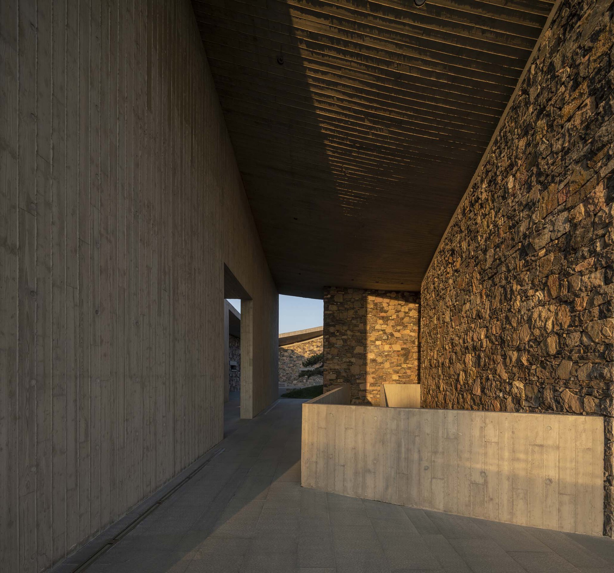 Interior corridor at dusk with exposed timber ceiling joists between concrete and stone walls