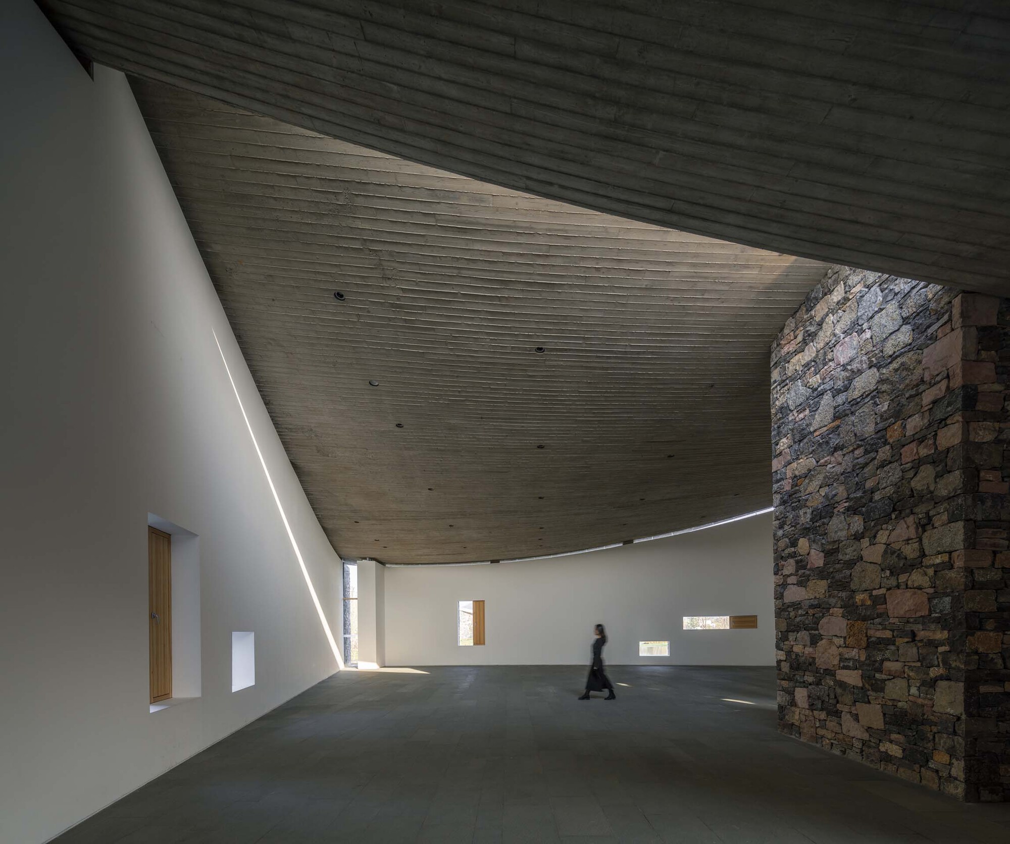 Interior hall with sloping timber ceiling and stone wall as a figure walks through sunlight