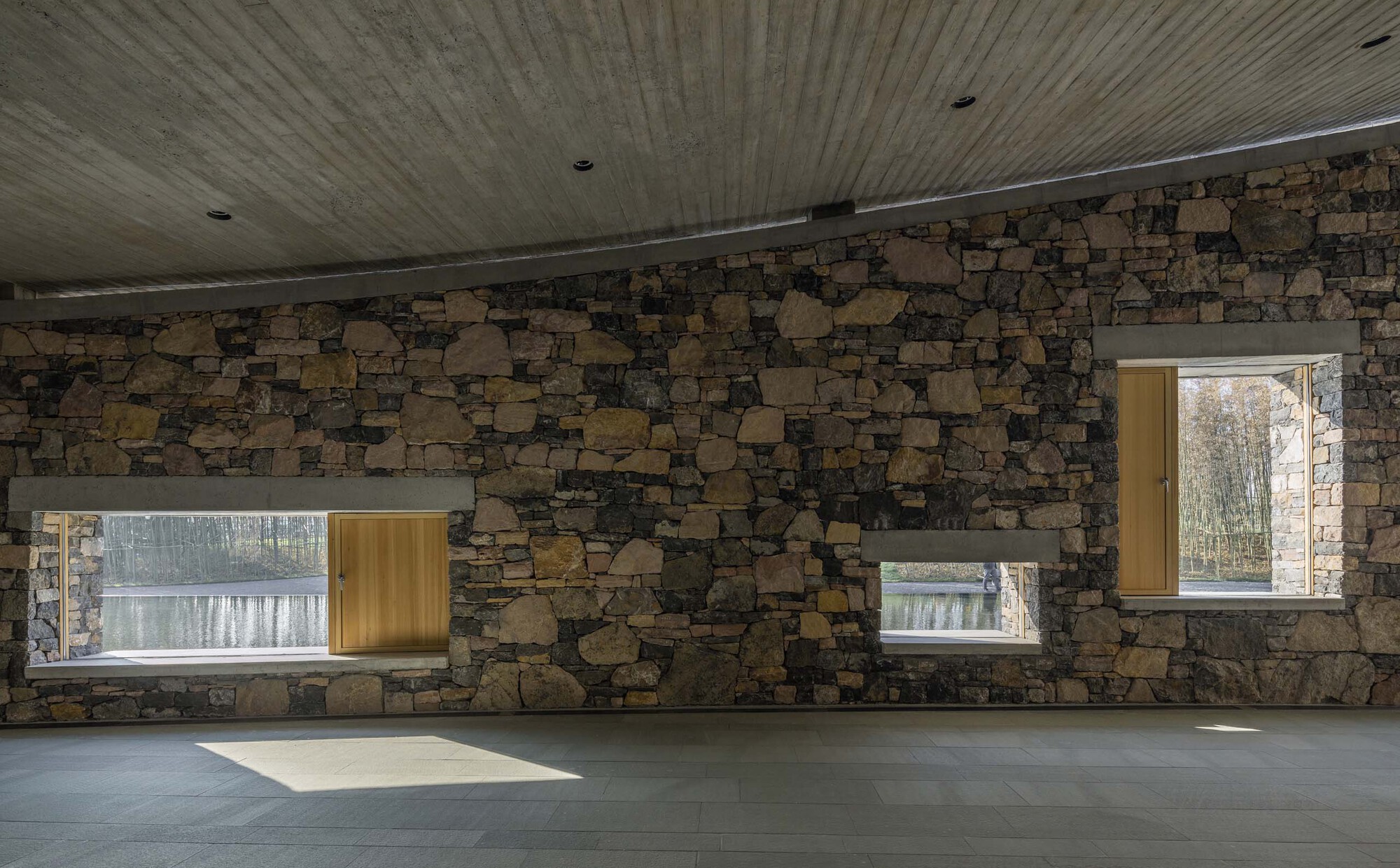 Fieldstone wall with timber-framed windows and clerestory slot admitting afternoon sunlight
